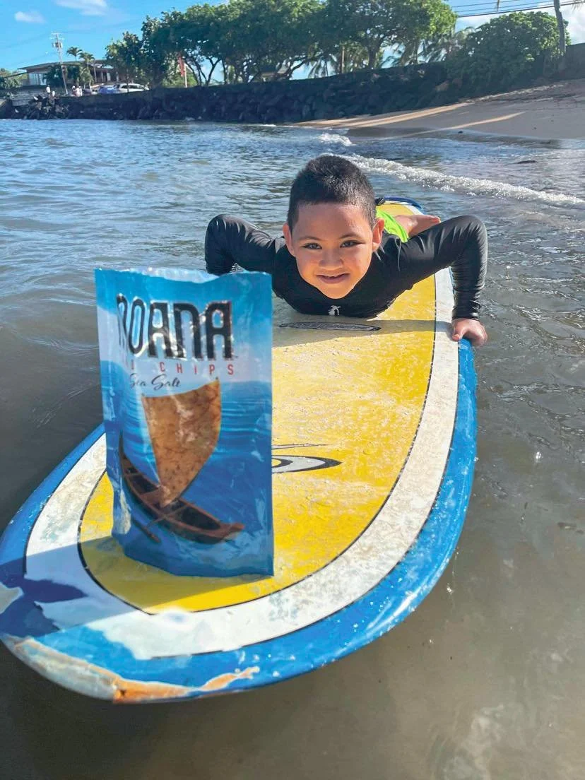 Child lying on a surfboard in shallow ocean water, smiling at the camera, with a bag of Moana taro chips chips placed in front.