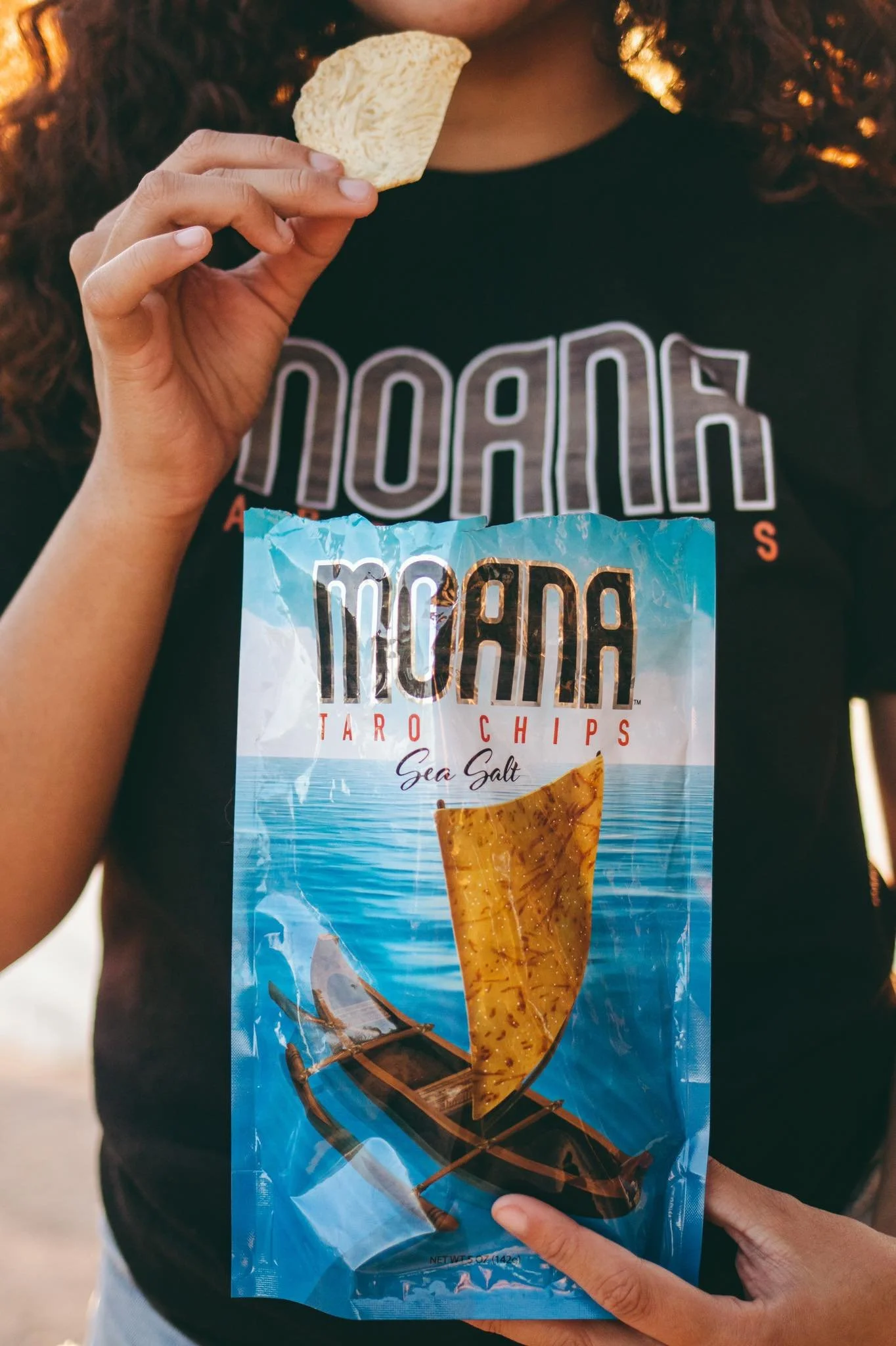 A person holding a bag of Taro Chips Sea Salt in front of their shirt with large text. The person has curly hair and is wearing a black shirt.