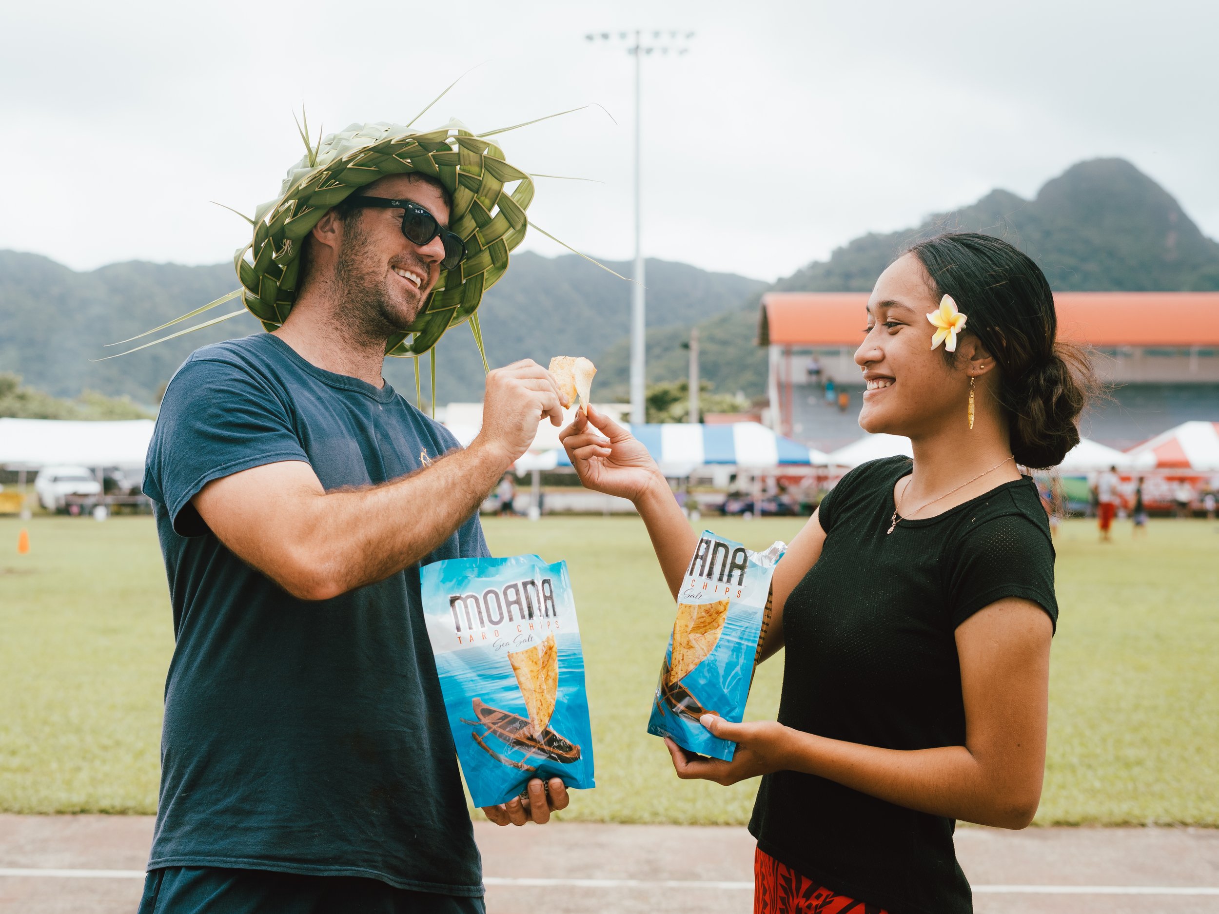 Two people exchanging a bag of moana taro chips outdoors, one wearing a woven hat and sunglasses, the other with a flower in her hair, at a park with mountains in the background.