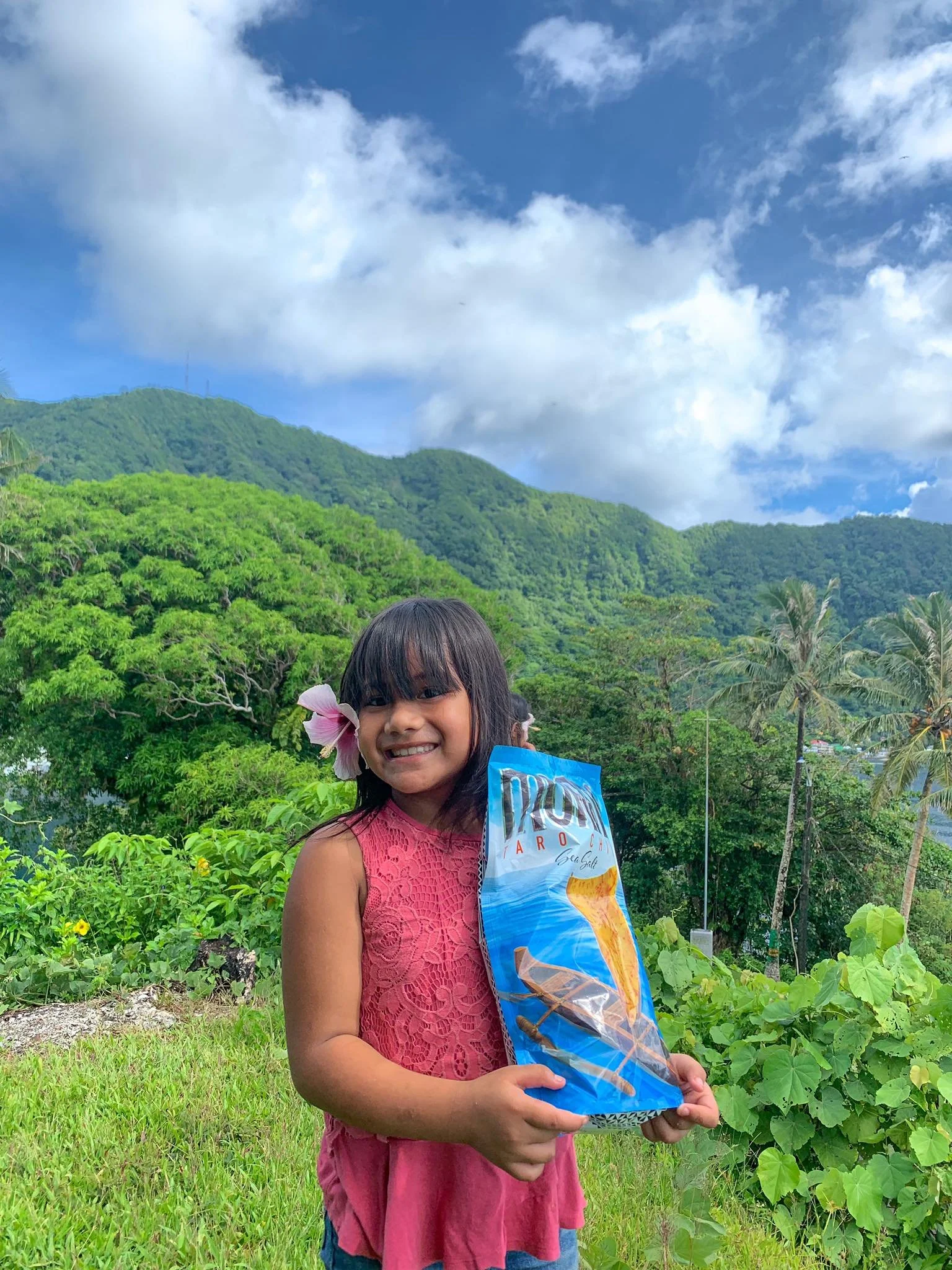 A young girl with black hair and bangs, wearing a pink sleeveless top and blue shorts, is outdoors in a lush green area holding a large bag  chips. She has a pink hibiscus flower behind her ear, and behind her is a background of vibrant green trees, bushes, and a hill with a partly cloudy blue sky.