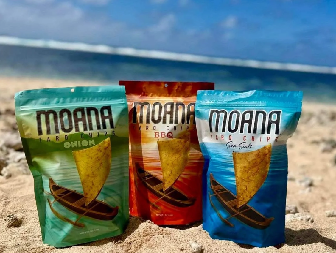 Three bags of Moana Taro Chips on the beach with the ocean in the background. The flavors are Onion, BBQ, and Sea Salt.