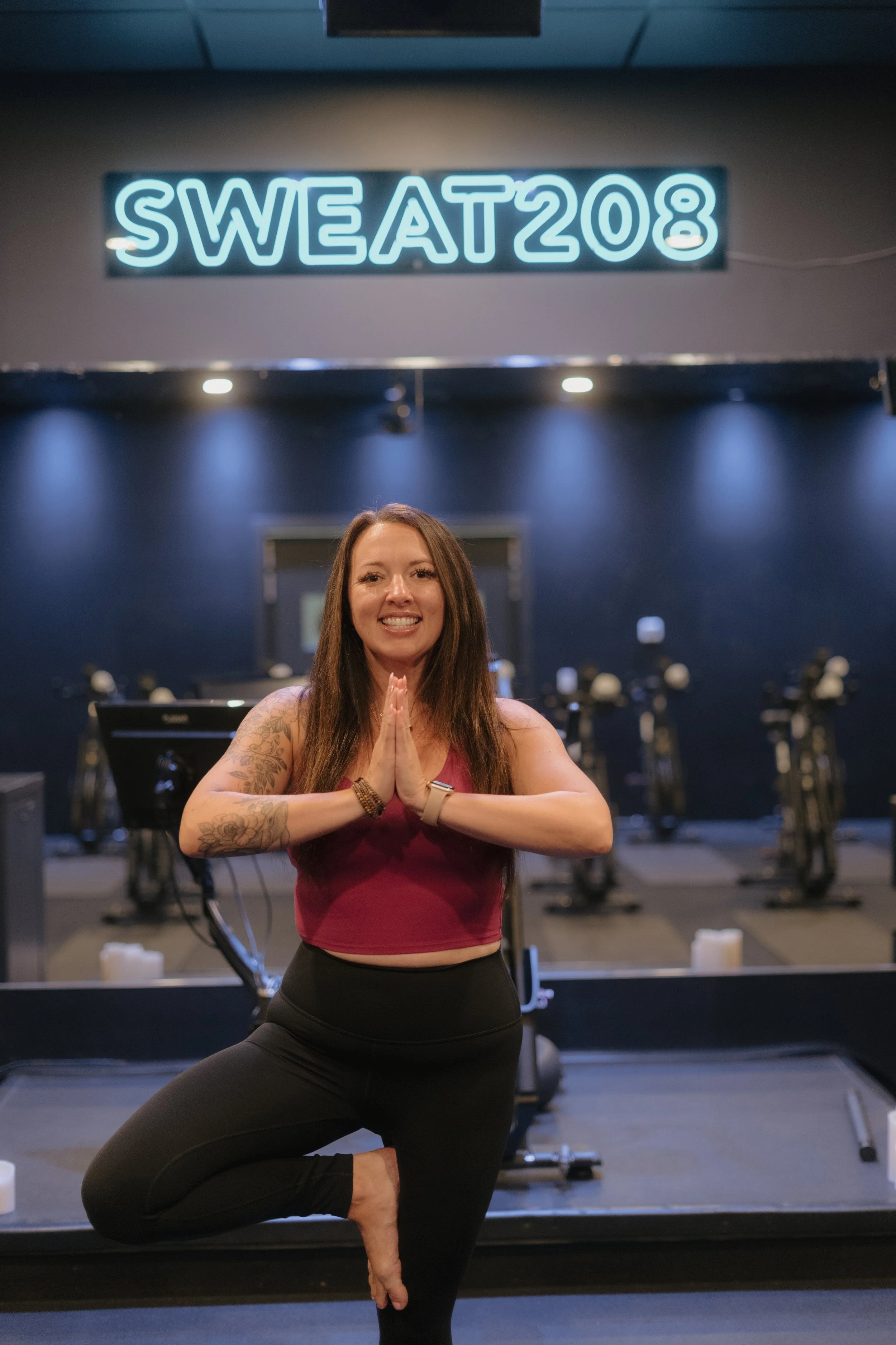 A woman practicing yoga or meditation in a gym, balancing on one leg in a tree pose with hands in prayer position, with a neon sign that reads 'SWEAT 208' in the background.