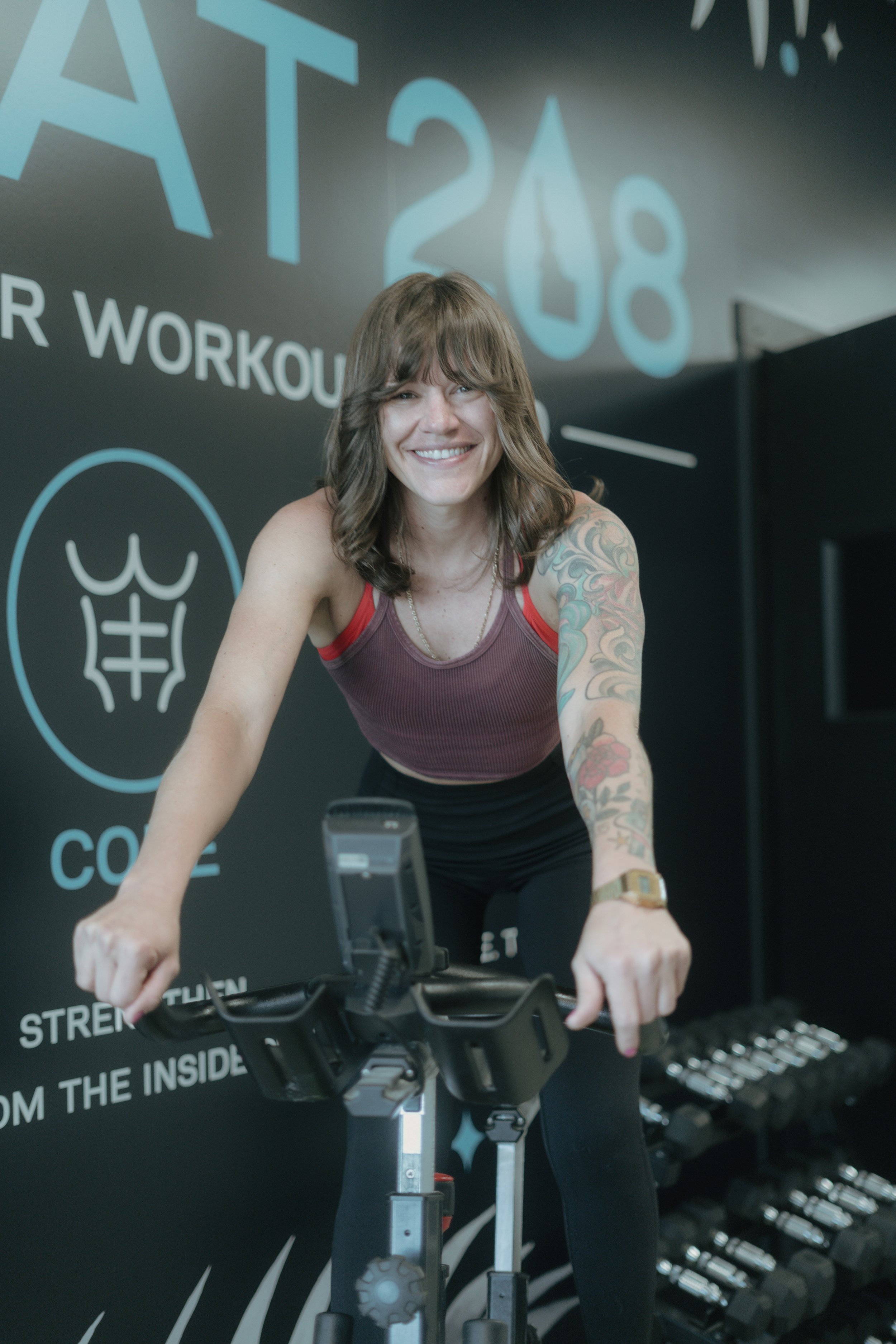 A woman with long brown hair, wearing a red tank top, smiling and exercising on a stationary bike in a gym. She has tattoos on her left arm and is looking toward the camera. The background features motivational text and a graphic of dumbbells.