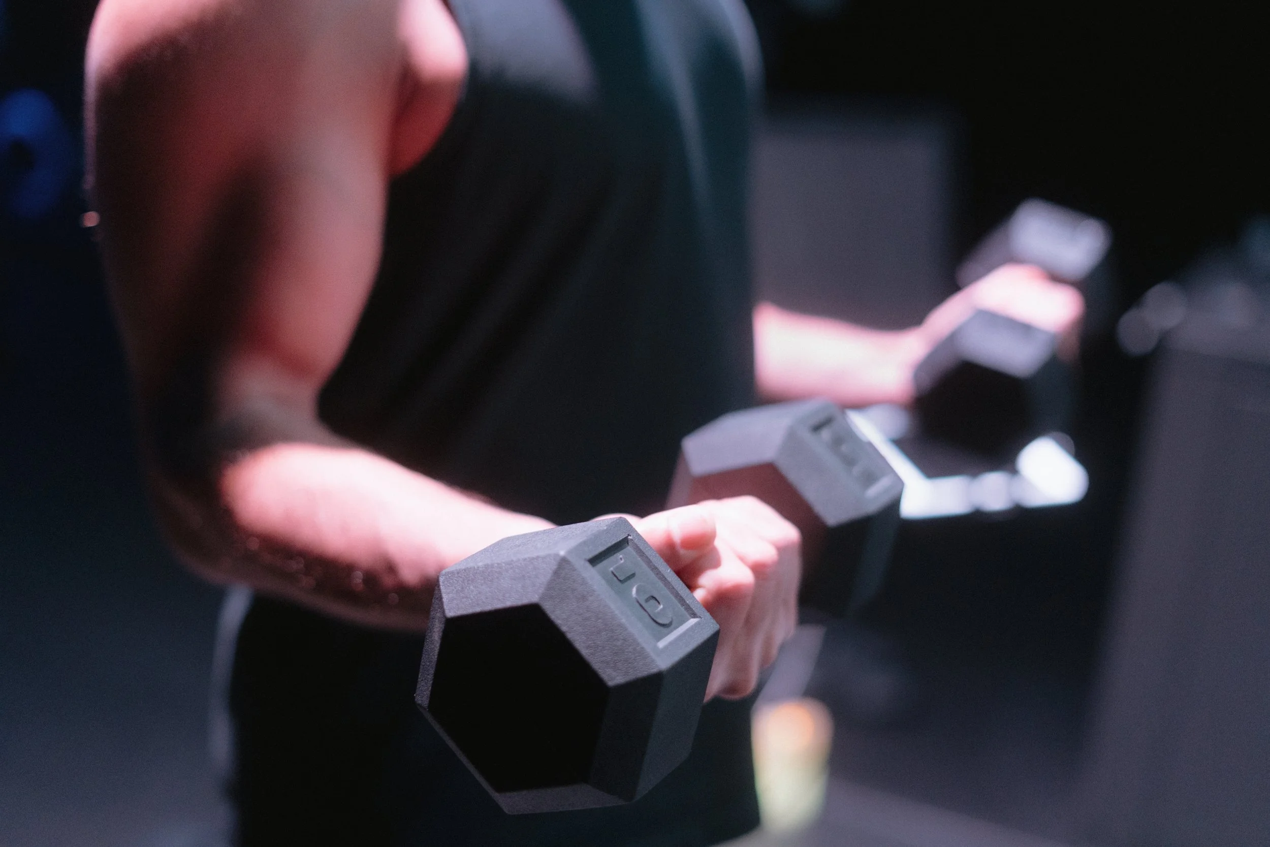 Close-up of a person's hand gripping a dumbbell with hexagonal weights, with a person in the background using a mobile device.