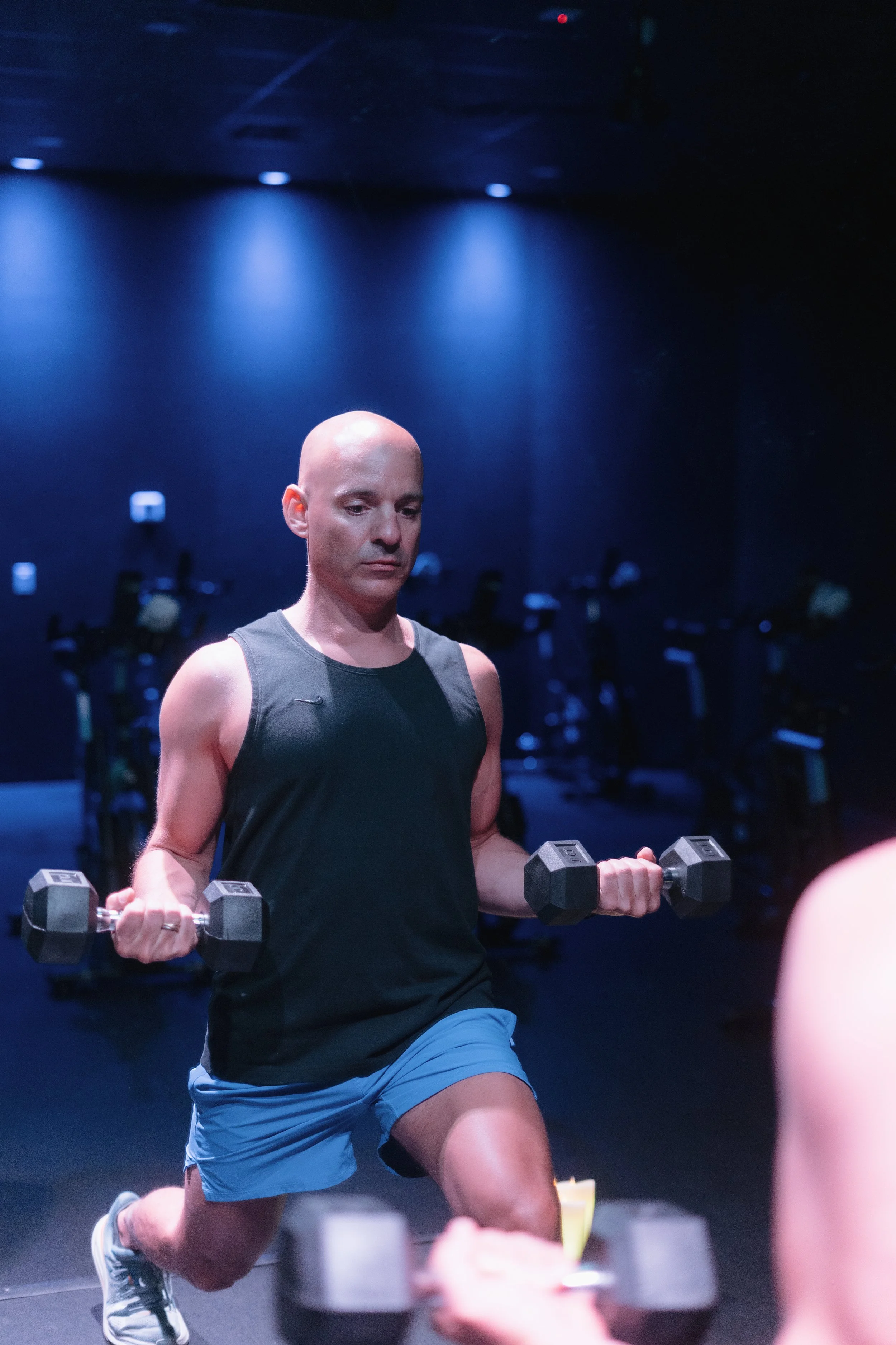 A man with a bald head and light skin is exercising in a gym, holding dumbbells and lifting weights while kneeling on one knee. He is wearing a black tank top, blue shorts, and sneakers. The gym has dark walls with blue lighting, and other exercise equipment is visible in the background.