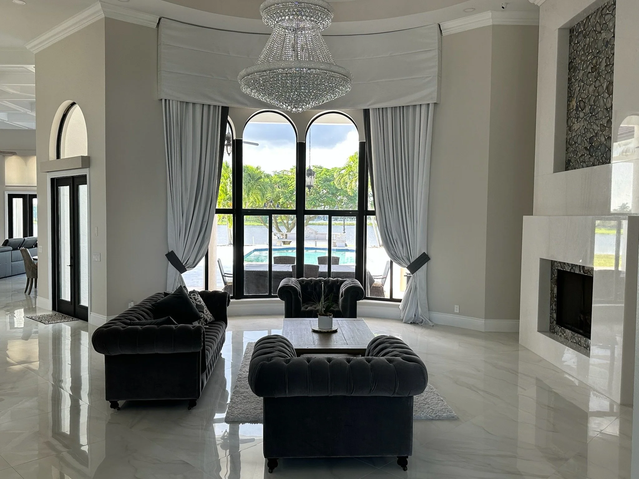 A spacious living room with cream-colored walls, a large chandelier hanging from the ceiling, and a view of a pool outside through floor-to-ceiling windows. The room is decorated with black velvet sofas and a white coffee table.