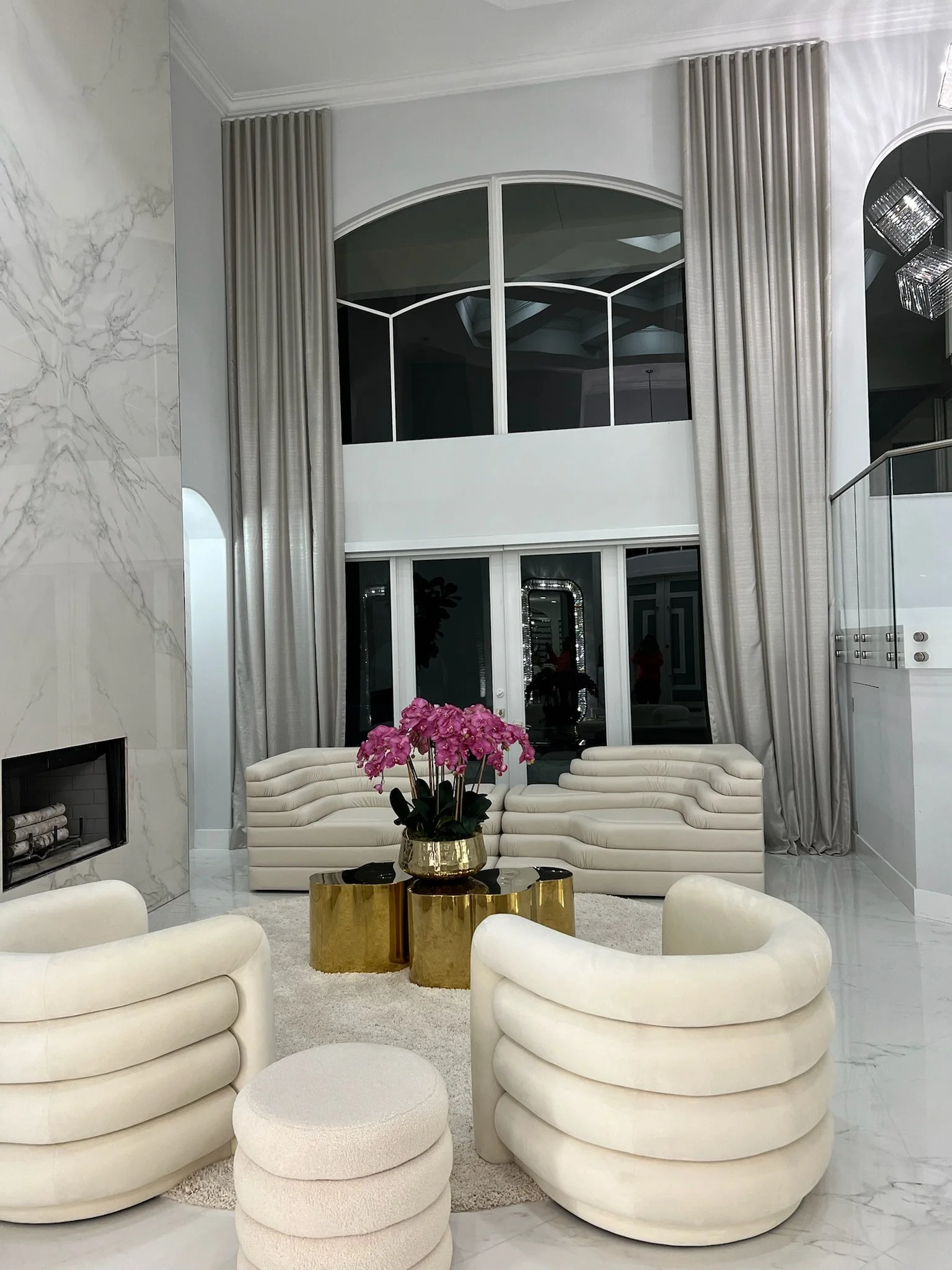 Elegant living room featuring a white curved sofa, matching armchairs, a floral arrangement on a gold and marble coffee table, and marble flooring with tall curtains and large windows.