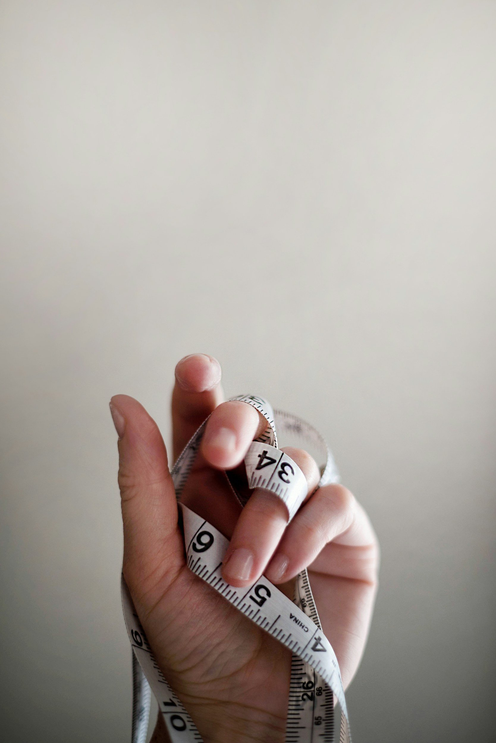 A person holding a white measuring tape with black numbers, against a plain light-colored background.