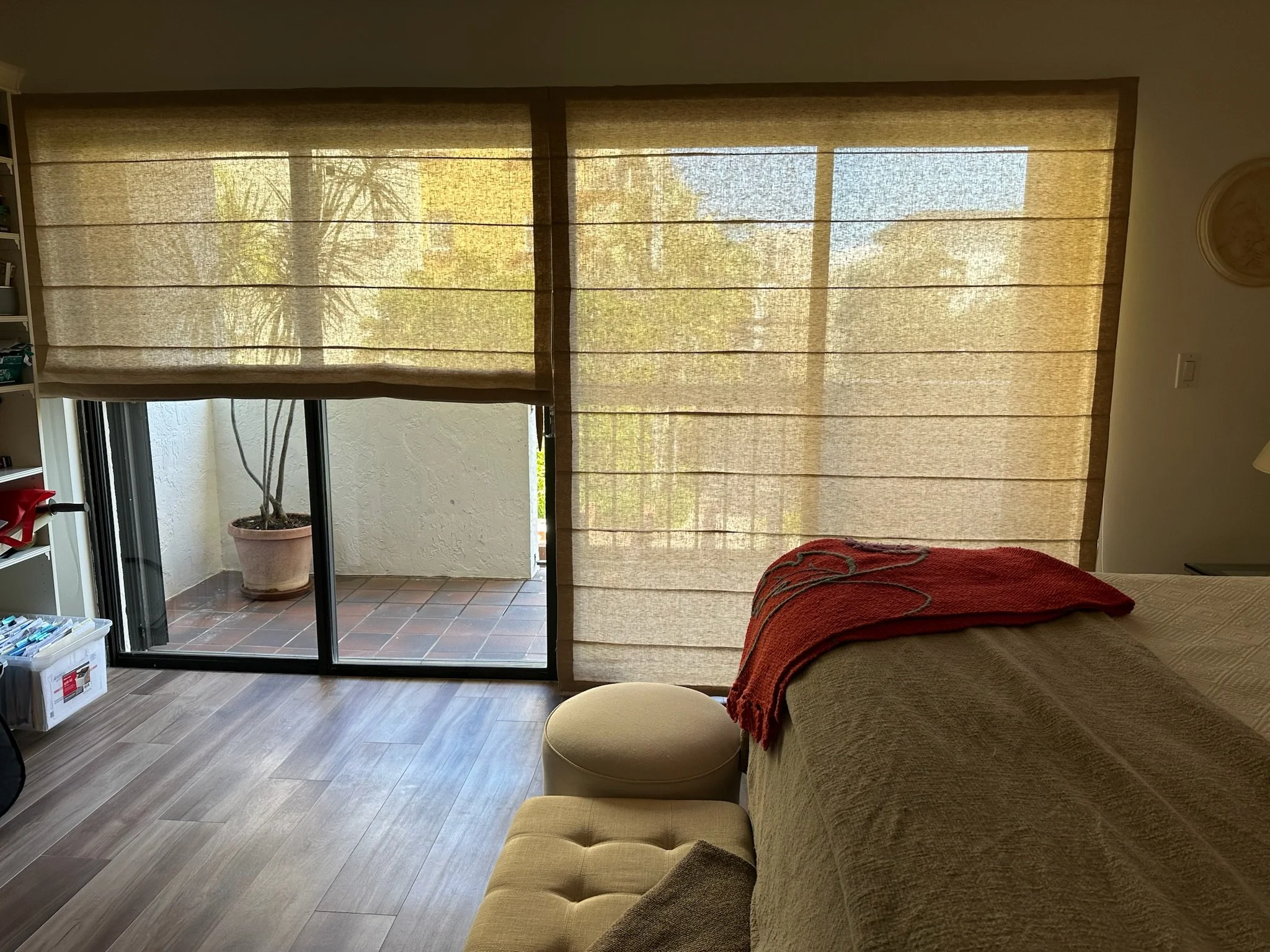 Living room with beige curtains covering sliding glass door, a potted plant outside, a bookcase on the left, a cozy couch with a blanket, a small ottoman, and a floor lamp.