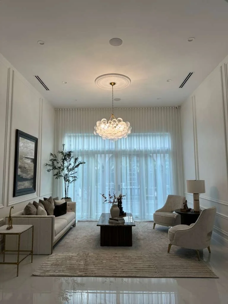 Bright living room with a modern chandelier, white walls with decorative molding, large window with sheer curtains, beige sofa and armchair, coffee table with decorative items, area rug, and a side table with a lamp.