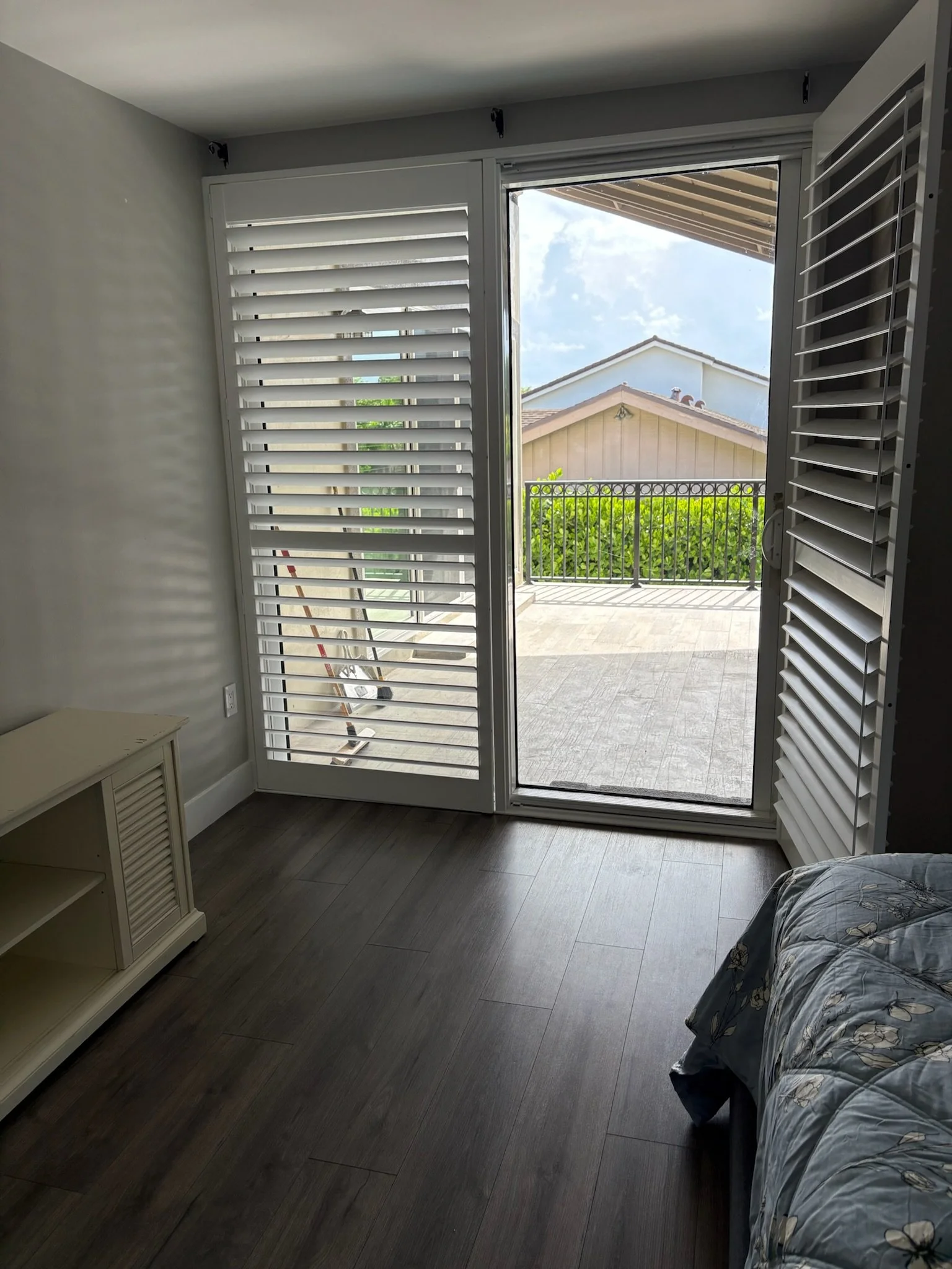 Room with sliding glass door leading to a patio, with white plantation shutters on either side, and a bed with a floral patterned blanket.