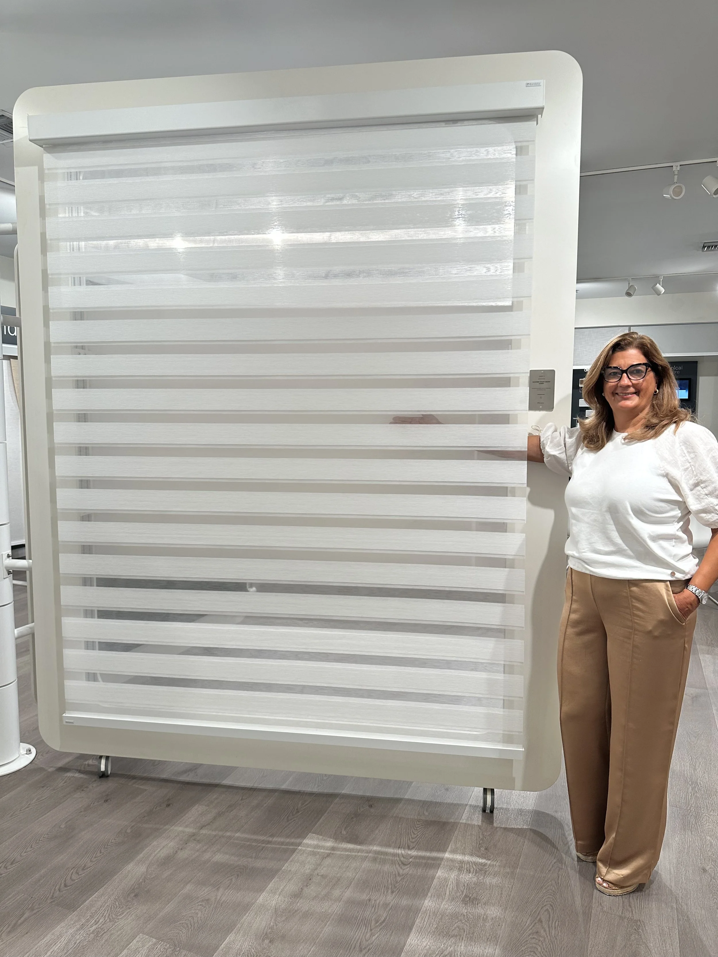 A woman with glasses and light brown hair standing next to a large window blind on a stand in an indoor modern setting.