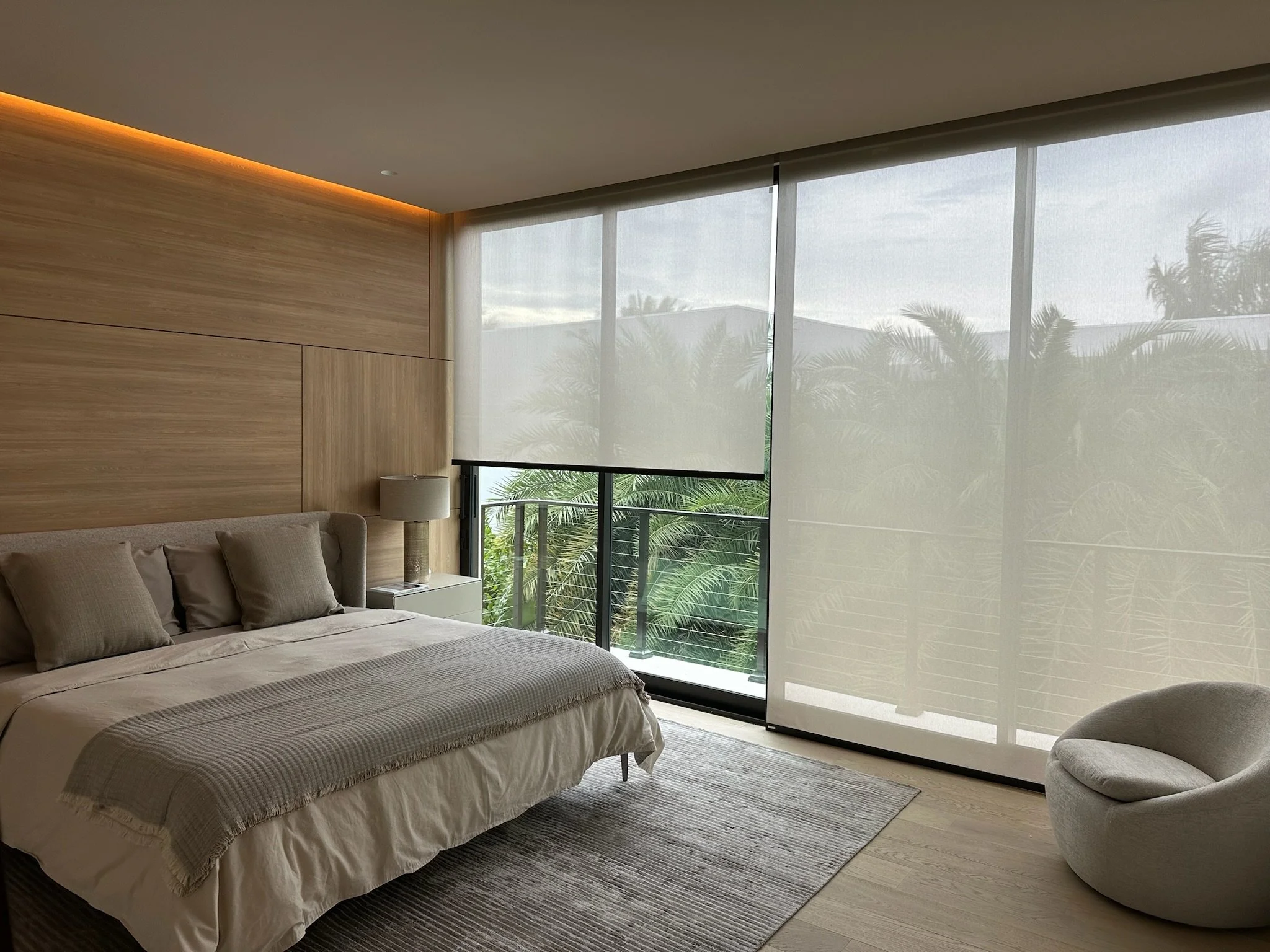 Modern bedroom with large window covered by sheer curtains, wooden wall accent, beige bed with pillows, and nightstand with a lamp. Tropical trees are visible outside.