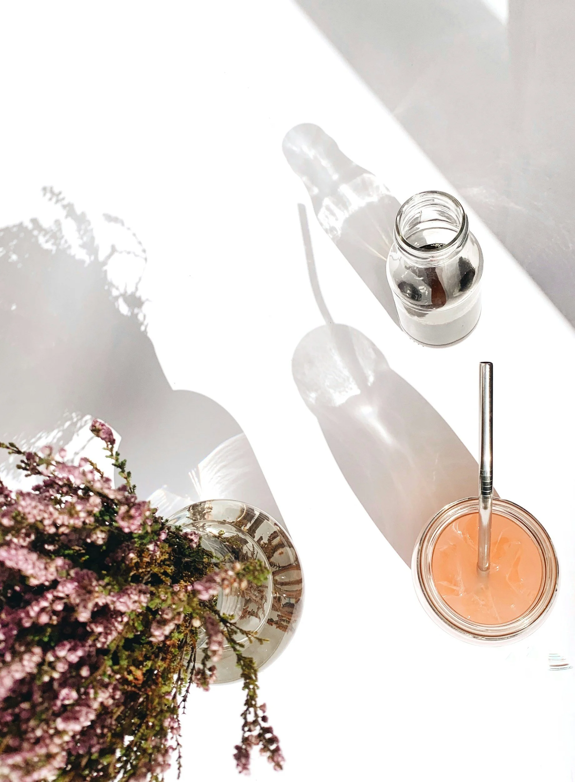 A top view of a table with a bouquet of pink flowers, a small glass bottle, a large glass containing a pink drink with a straw, and shadows cast by sunlight on a white surface.