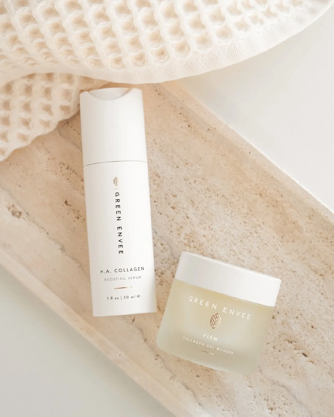 Two skincare products from Green Envee, a serum and a gel masque, placed on a beige textured surface with a cream knitted fabric and a white wall in the background.