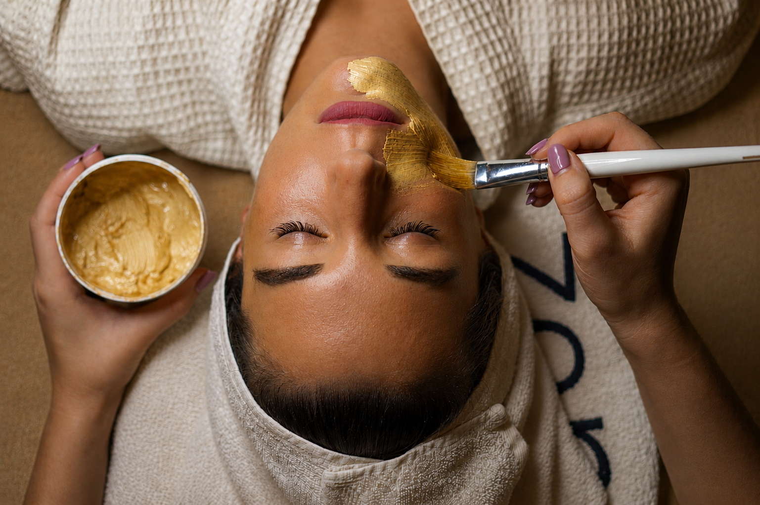 A woman is lying down with her eyes closed, receiving a facial treatment. A skincare therapist is applying a gold-colored mask to her face using a brush, and a container of the mask is held in her other hand.