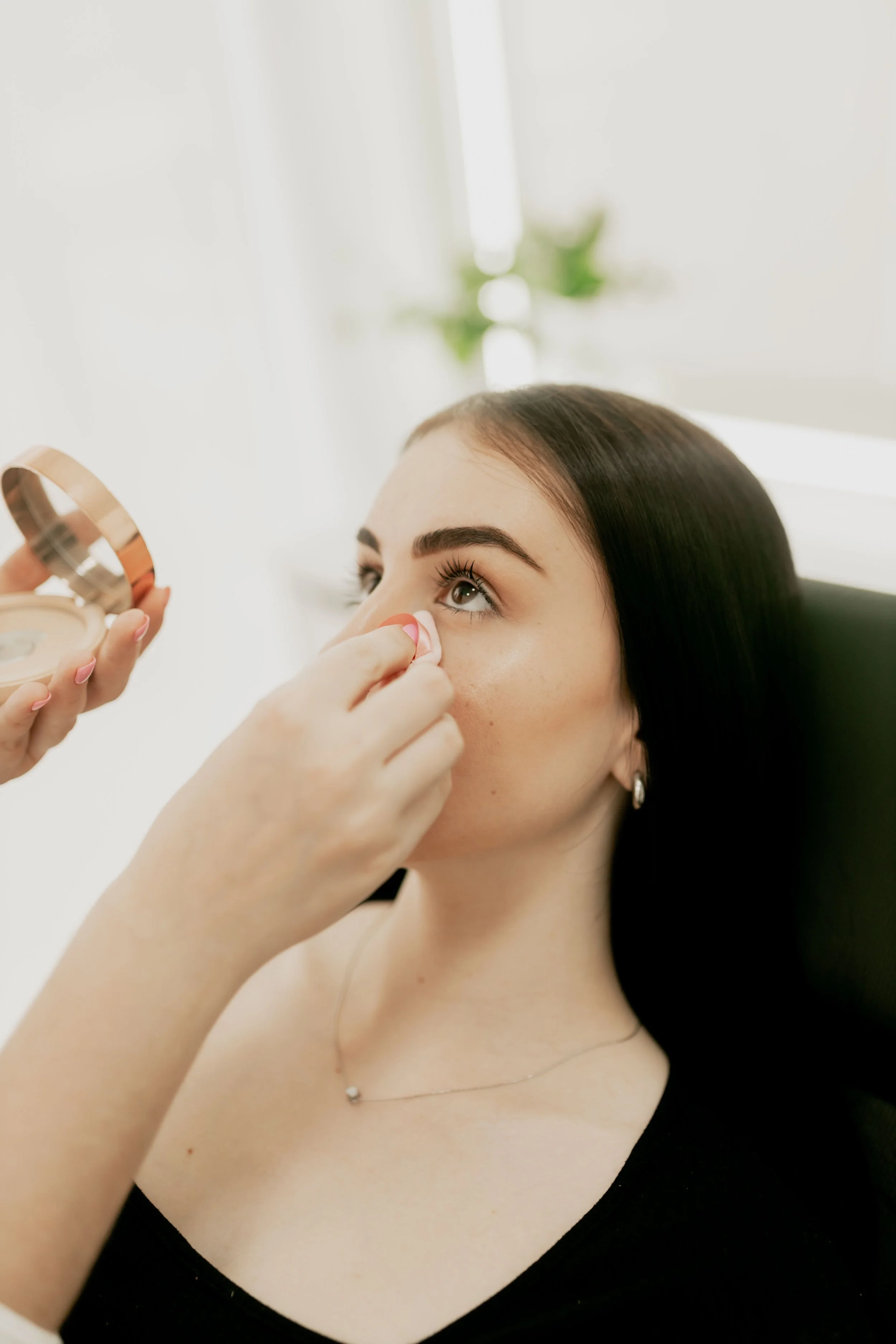 A woman with dark hair and earrings is applying makeup to her face with a compact mirror.