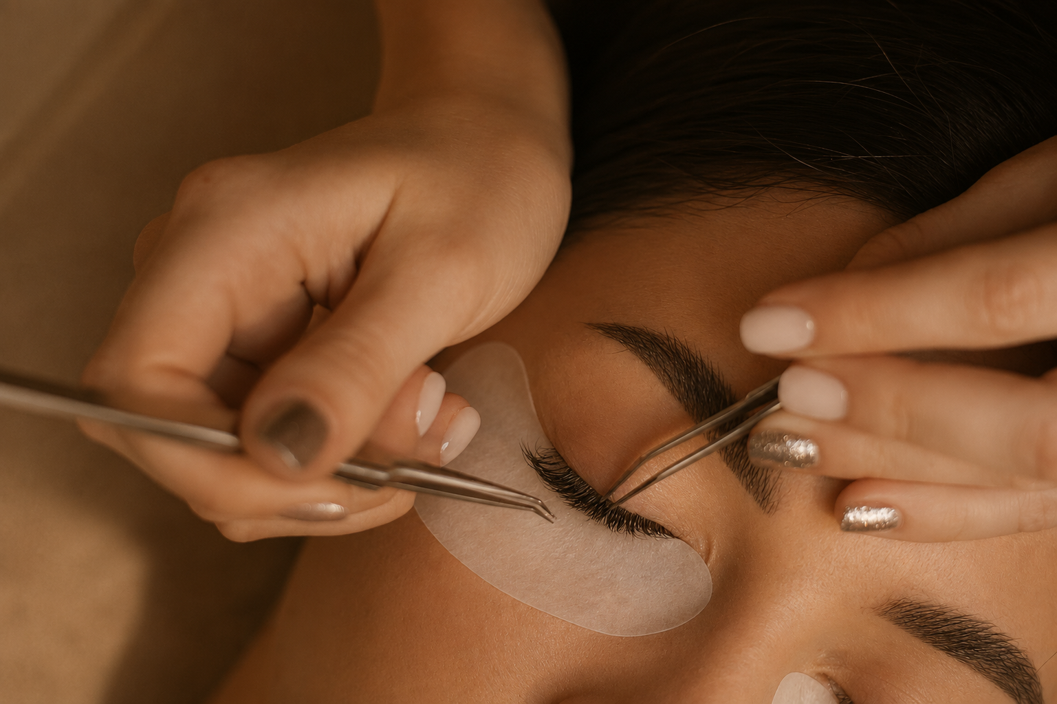 Close-up of a person having eyelash extension applied by a technician using tweezers, with eye pad under the eye for protection.