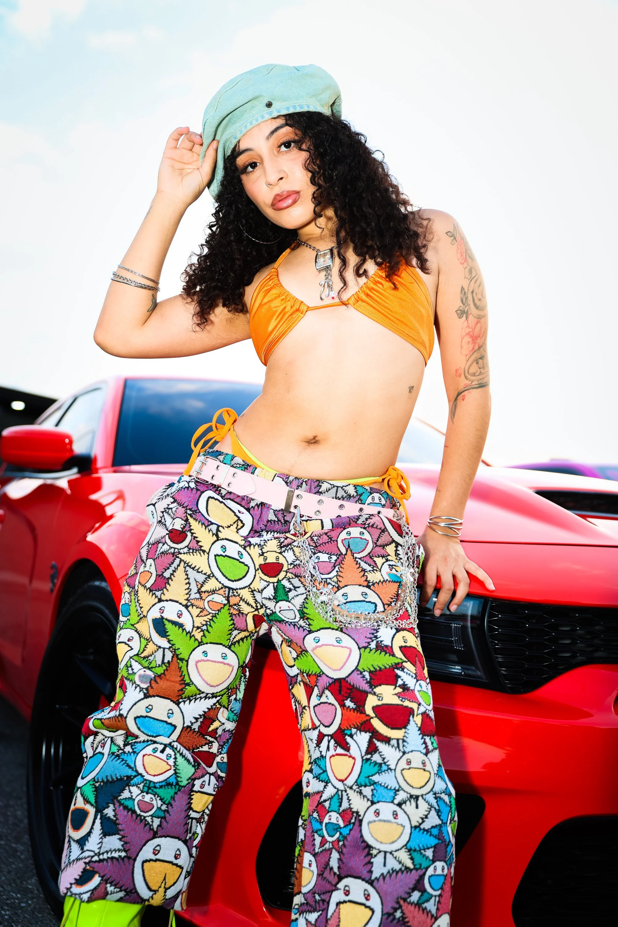 A woman with curly black hair wearing a green hat, orange top, colorful cartoon character and leaf print pants, and jewelry posing in front of a red sports car.