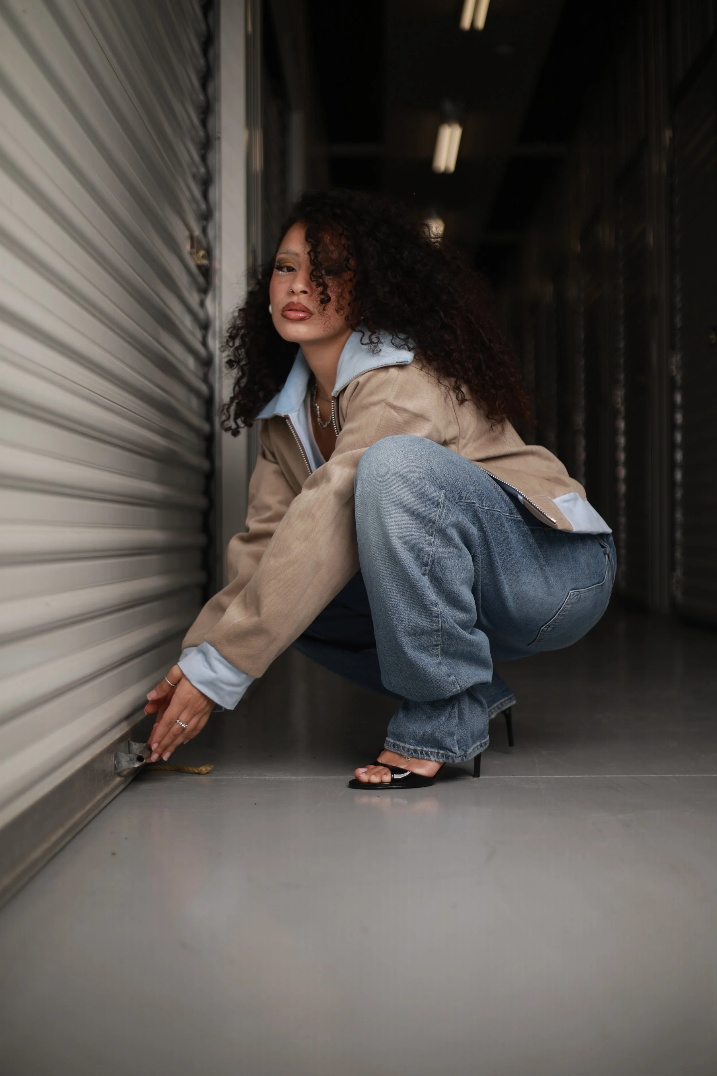 A woman with curly hair squatting and unlocking a storage unit in an industrial-style hallway.