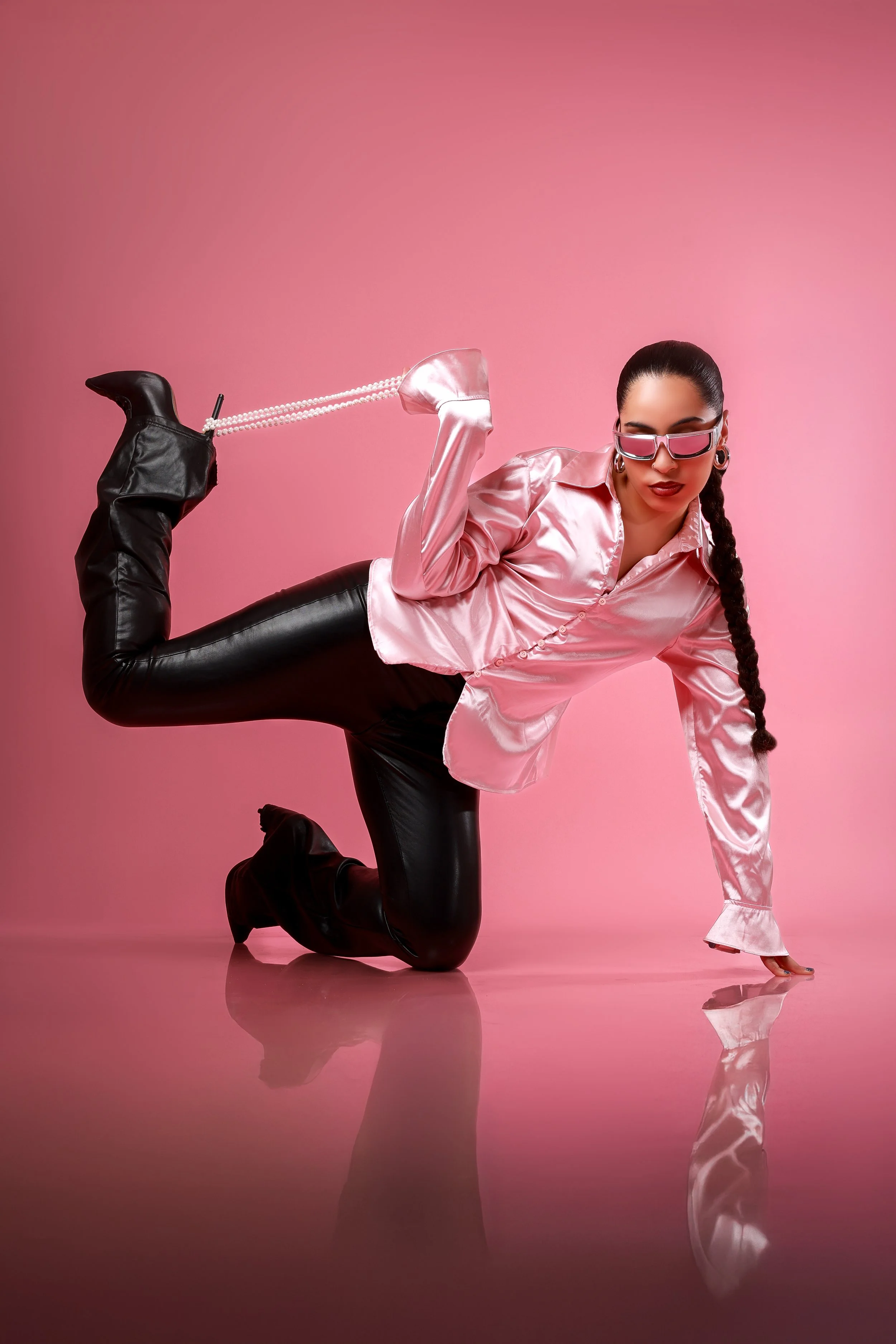 A woman in a pink satin blouse, black leather pants, and black boots is performing a dance move against a pink background, with her hand on the floor and her leg lifted, holding a pearl necklace.