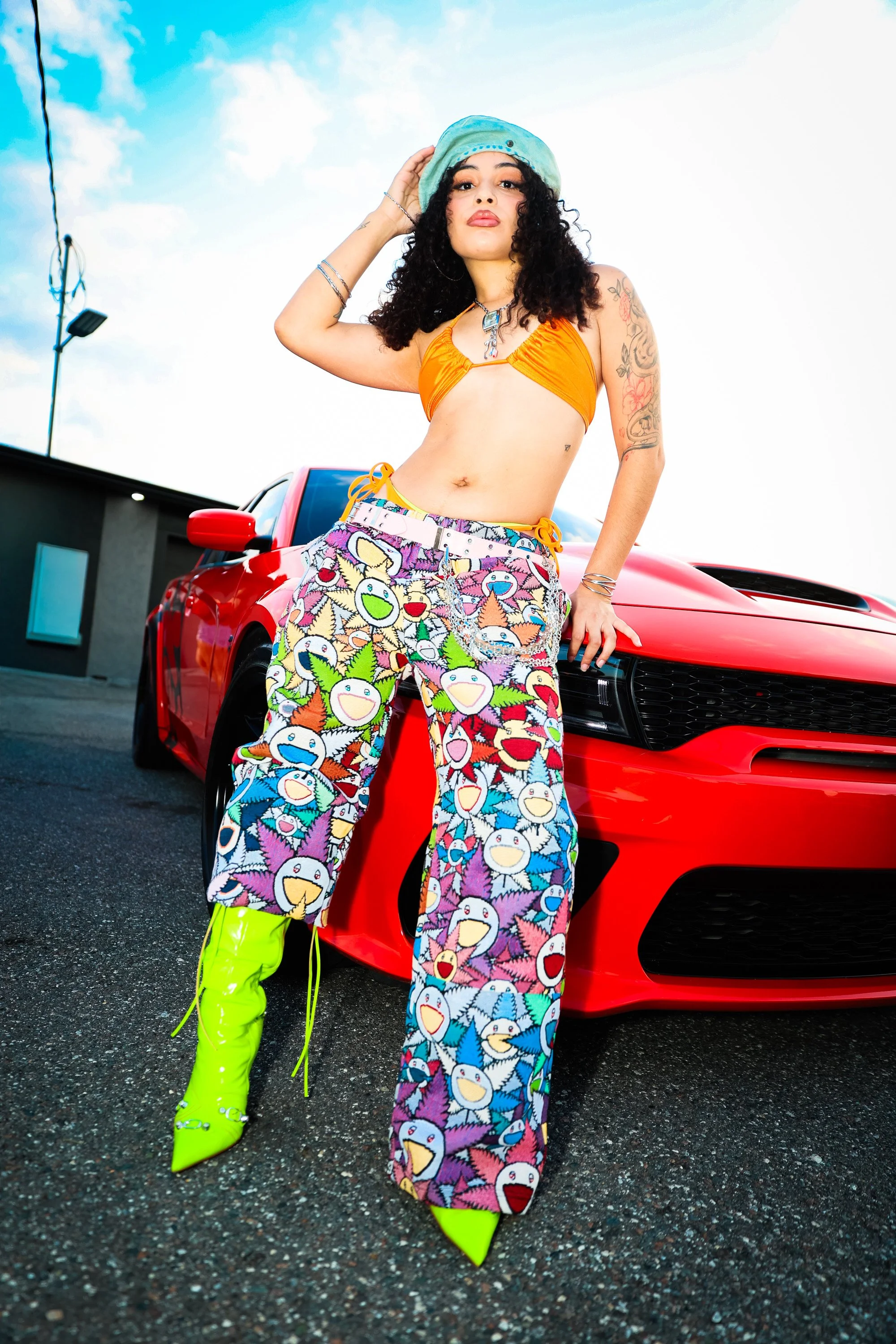 A woman with curly black hair, tattoos on her arm, and wearing bright green boots, standing in front of a red sports car, holding a light blue bucket hat, with a blue sky and a studio light in the background.