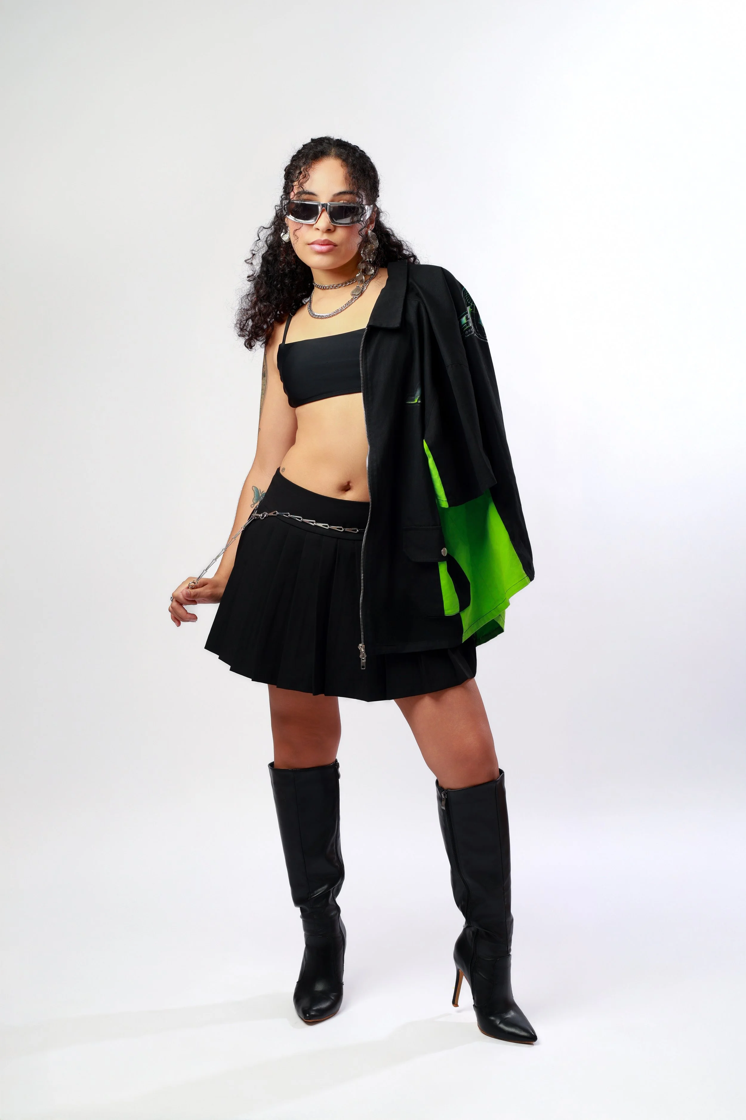 Woman with curly hair wearing sunglasses, layered necklaces, black crop top, black pleated skirt, a black jacket with green accents draped over her shoulder, and black knee-high boots, standing against a plain white background.