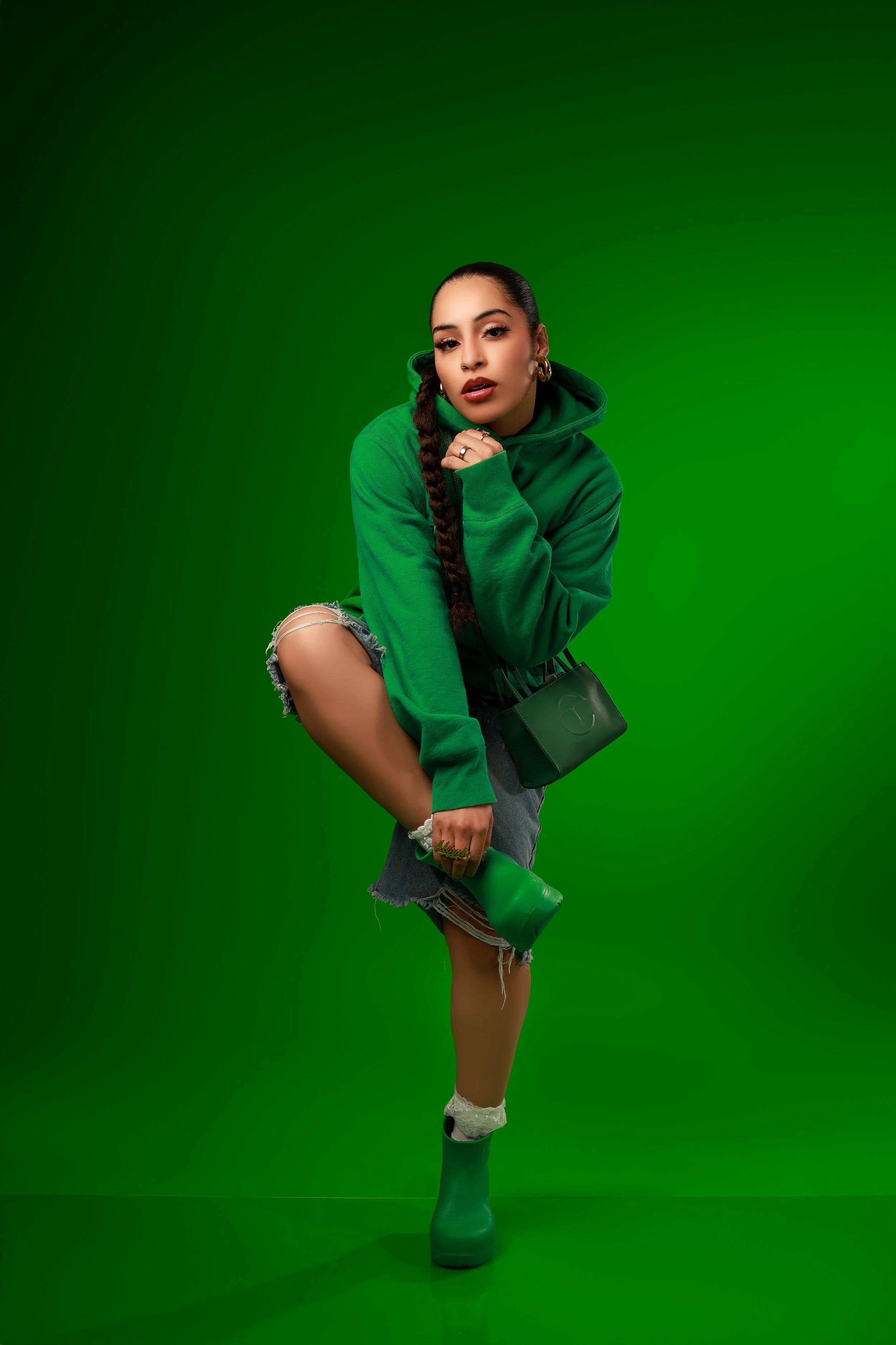 Young woman in green hoodie, ripped denim shorts, and green rain boots holding a green boot and a green purse, posing against a green background.