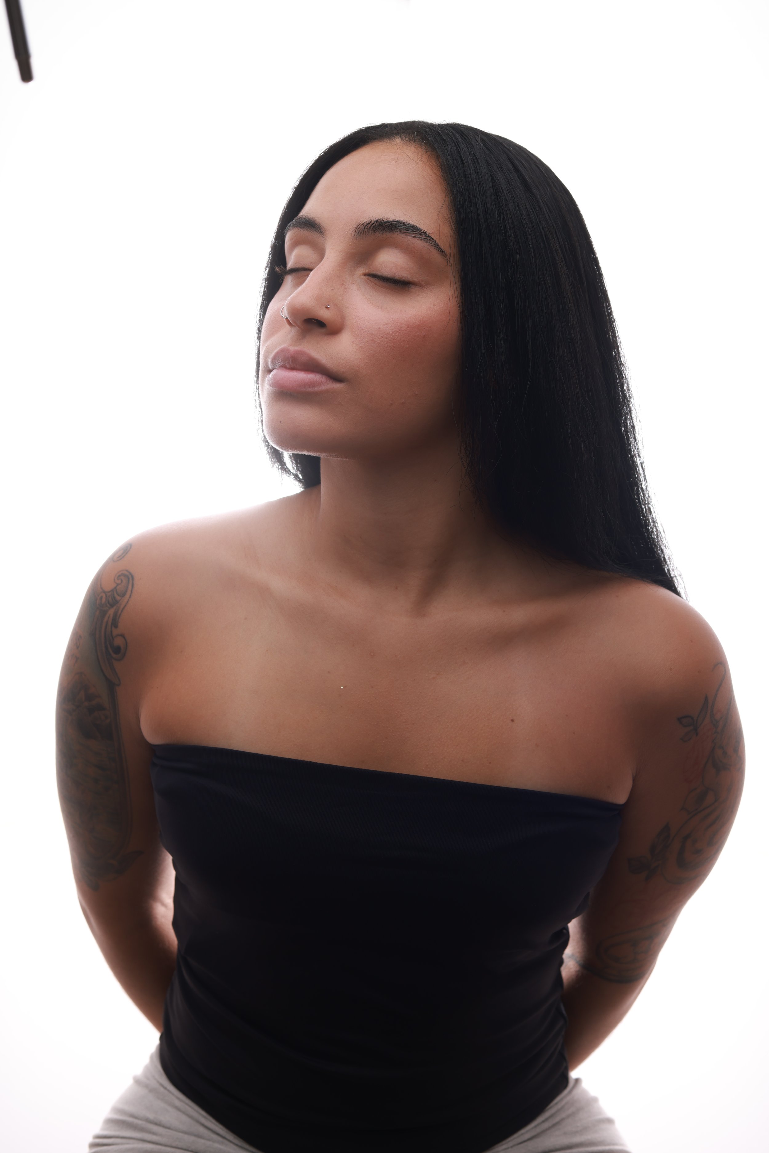 A woman with long black hair and tattoos on her arms, wearing a strapless black top with her eyes closed, against a plain white background.