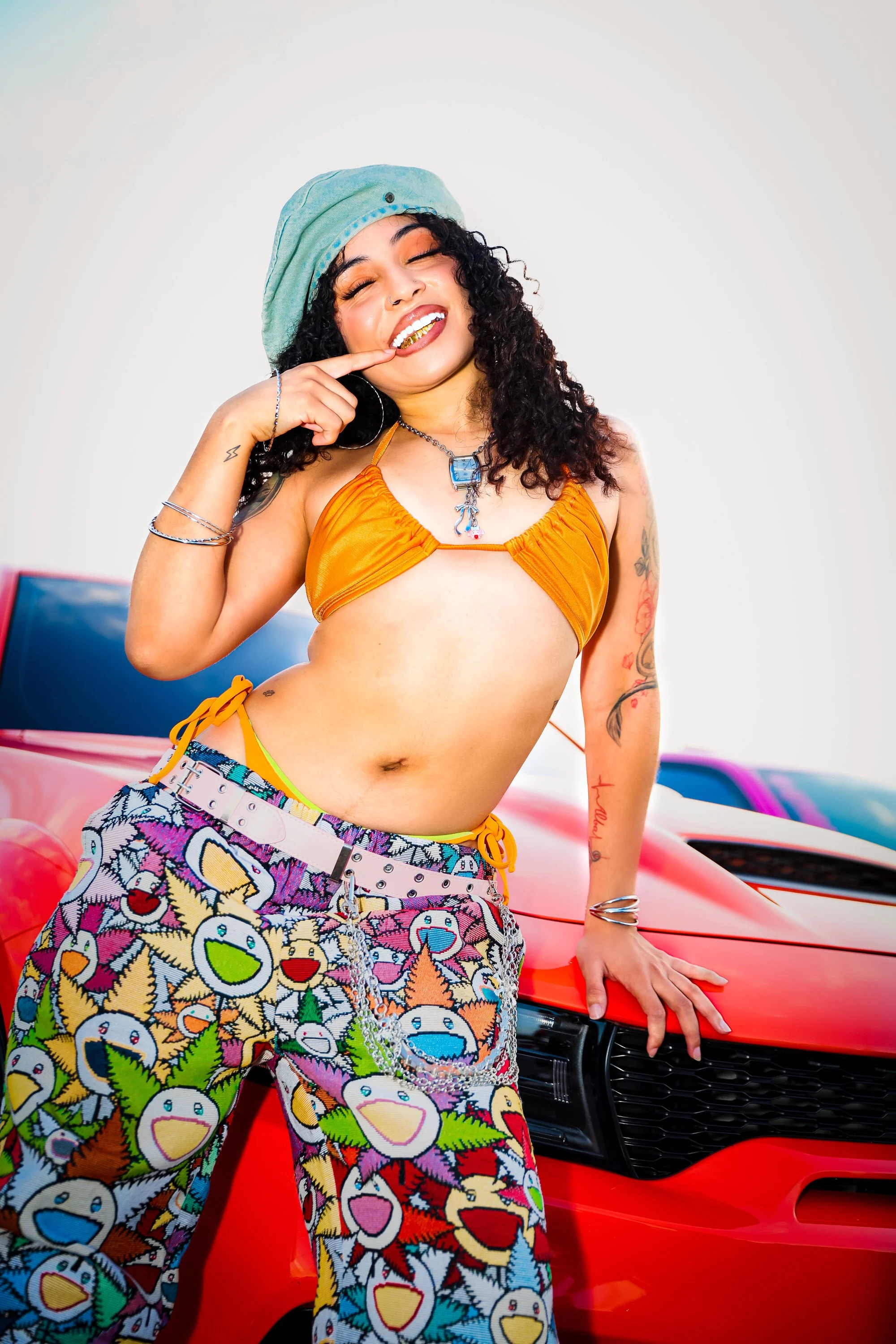 A woman with curly black hair wearing a green headscarf, gold grillz, orange bikini top, colorful patterned pants, and jewelry, standing beside a red sports car, smiling and touching her lip with one finger.