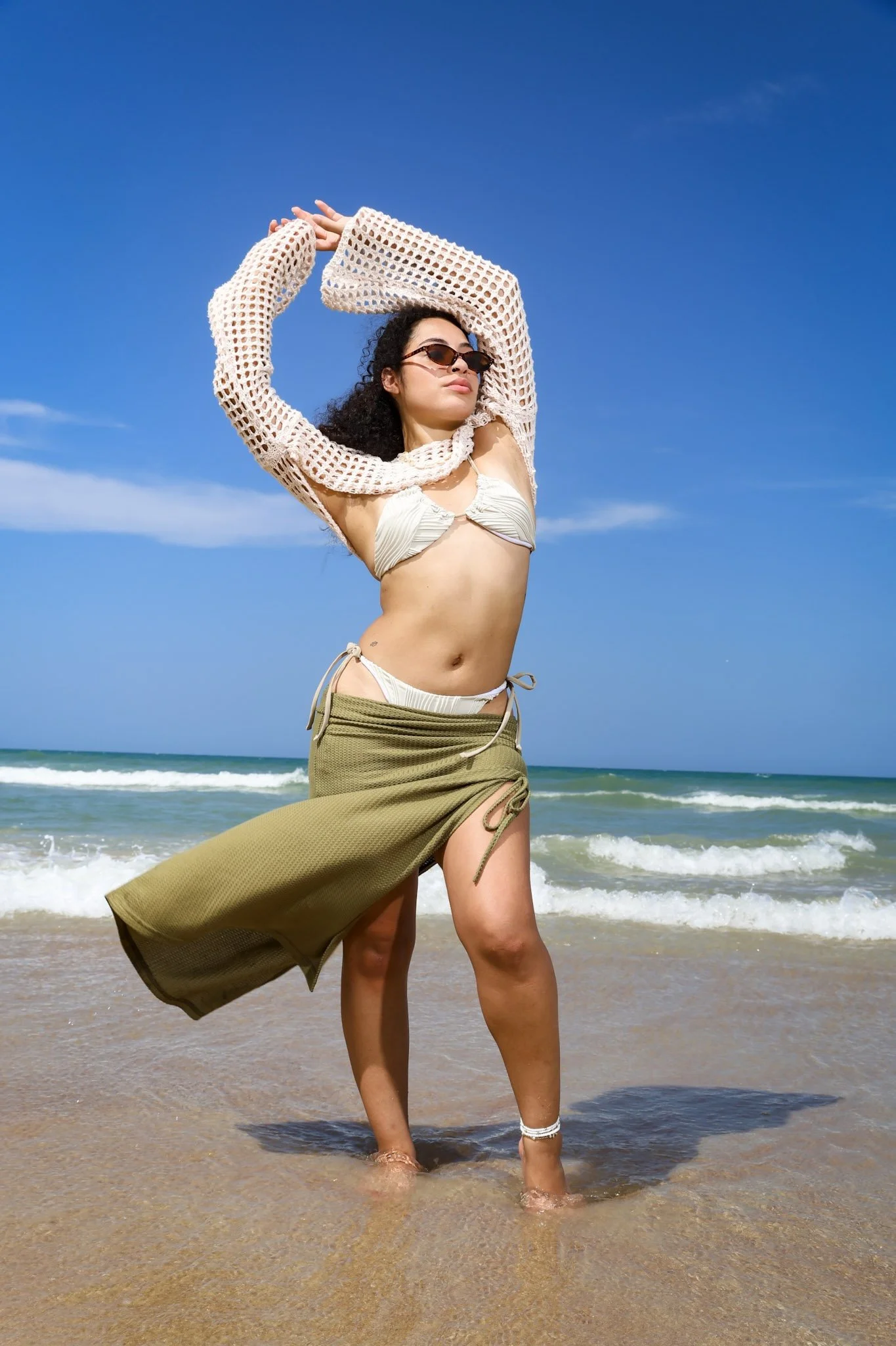 Woman standing in the ocean waves on a sandy beach, wearing a bikini, sunglasses, and a mesh cover-up with an olive green skirt.