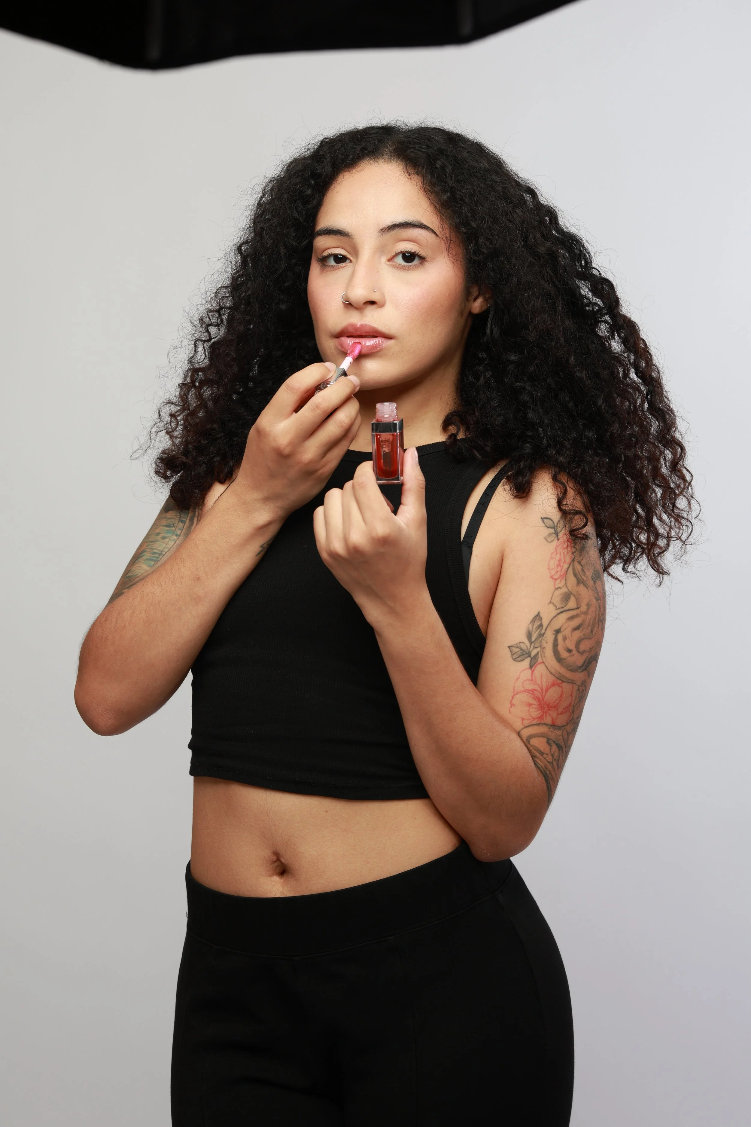 A woman with curly dark hair applies pink lipstick using a small brush, holding a lipstick tube, standing against a plain gray background.