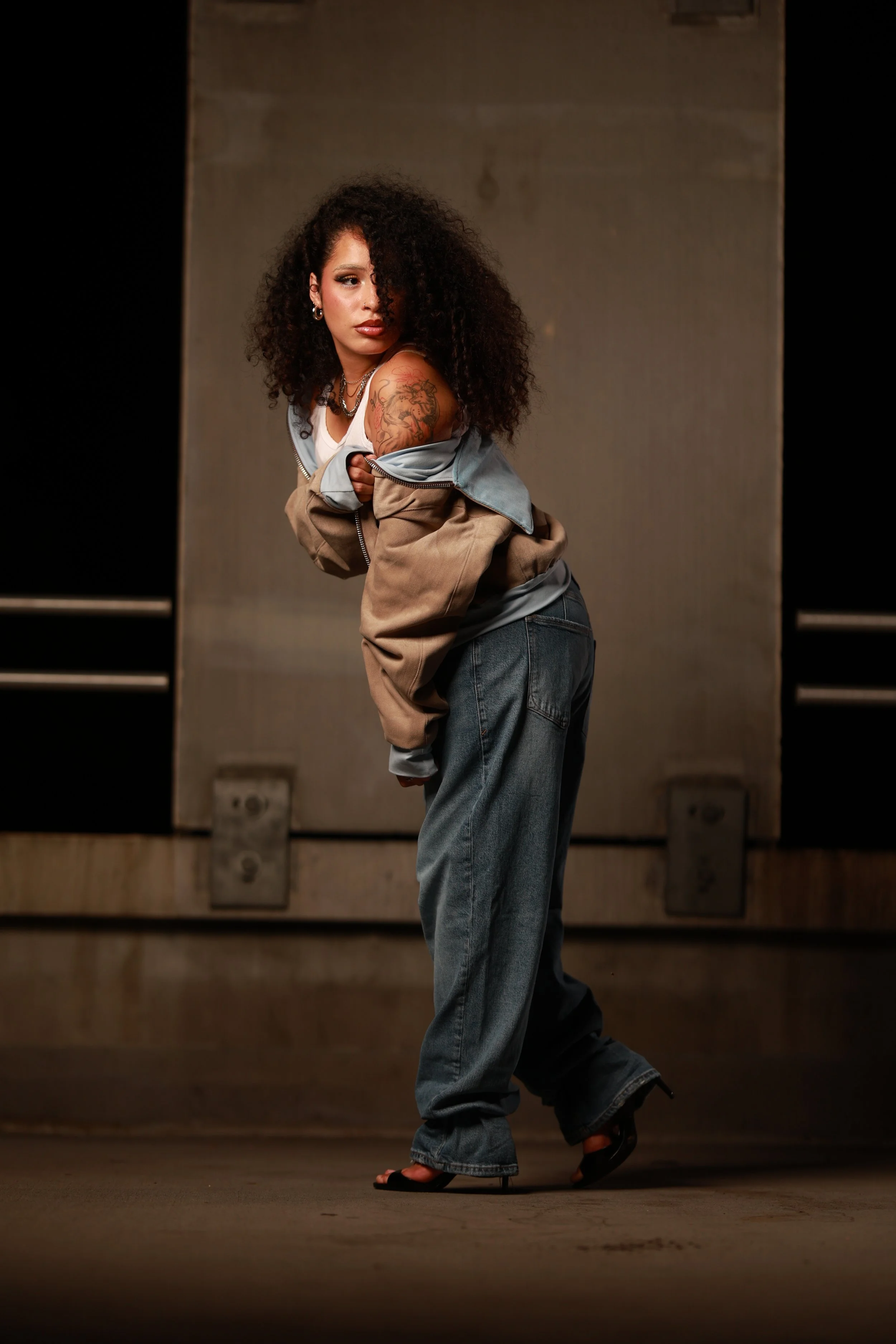 A woman with curly hair and tattoos on her arm stands against a plain background, wearing a jacket off her shoulders, jeans, and high heels.