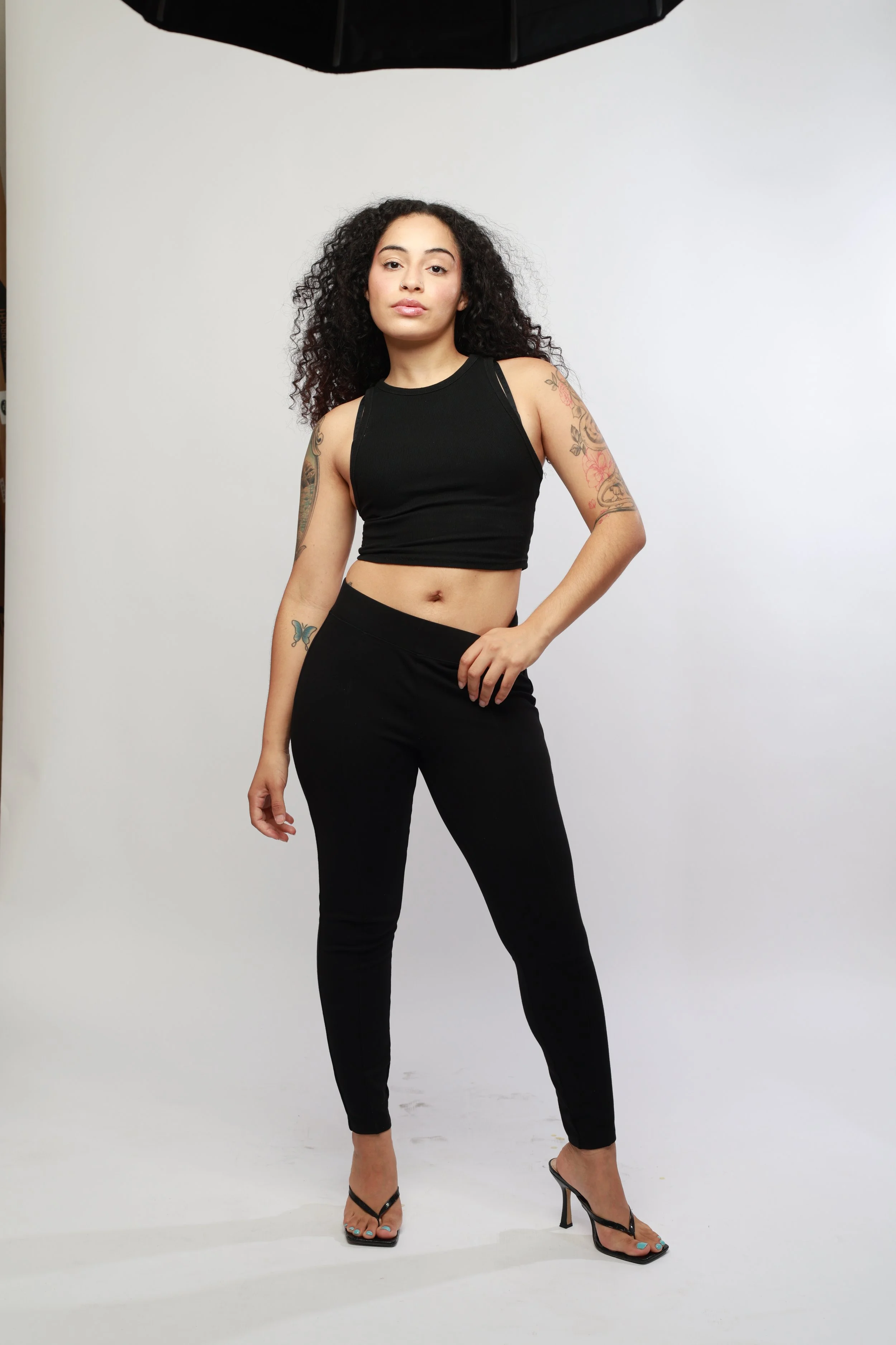 A woman with curly dark hair, tattoos on her arms, wearing a black sleeveless crop top, black pants, and high-heeled sandals, standing in front of a plain white background.