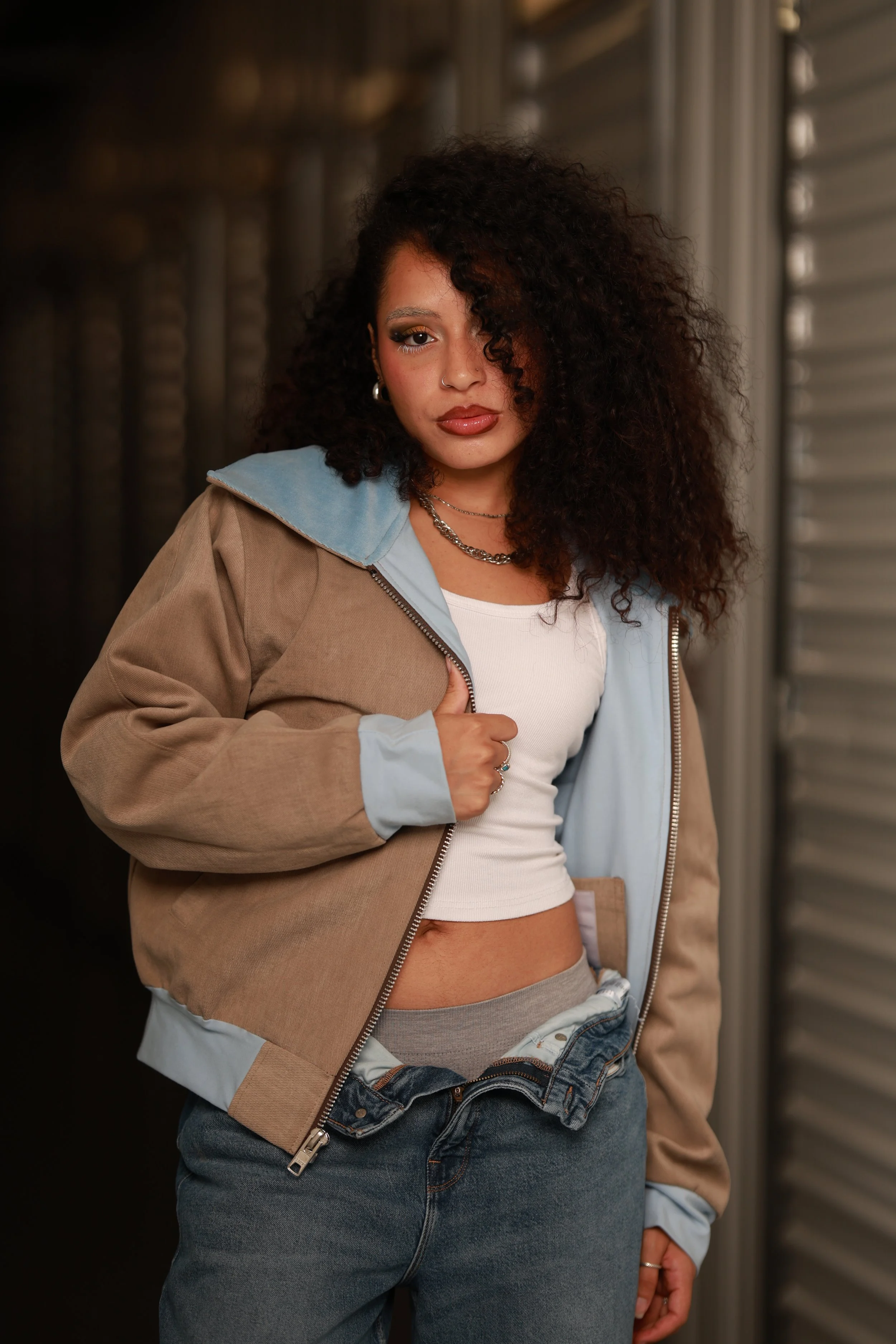 A woman with curly dark hair wearing a beige and light blue jacket, white crop top, and jeans, standing in front of a metal wall.