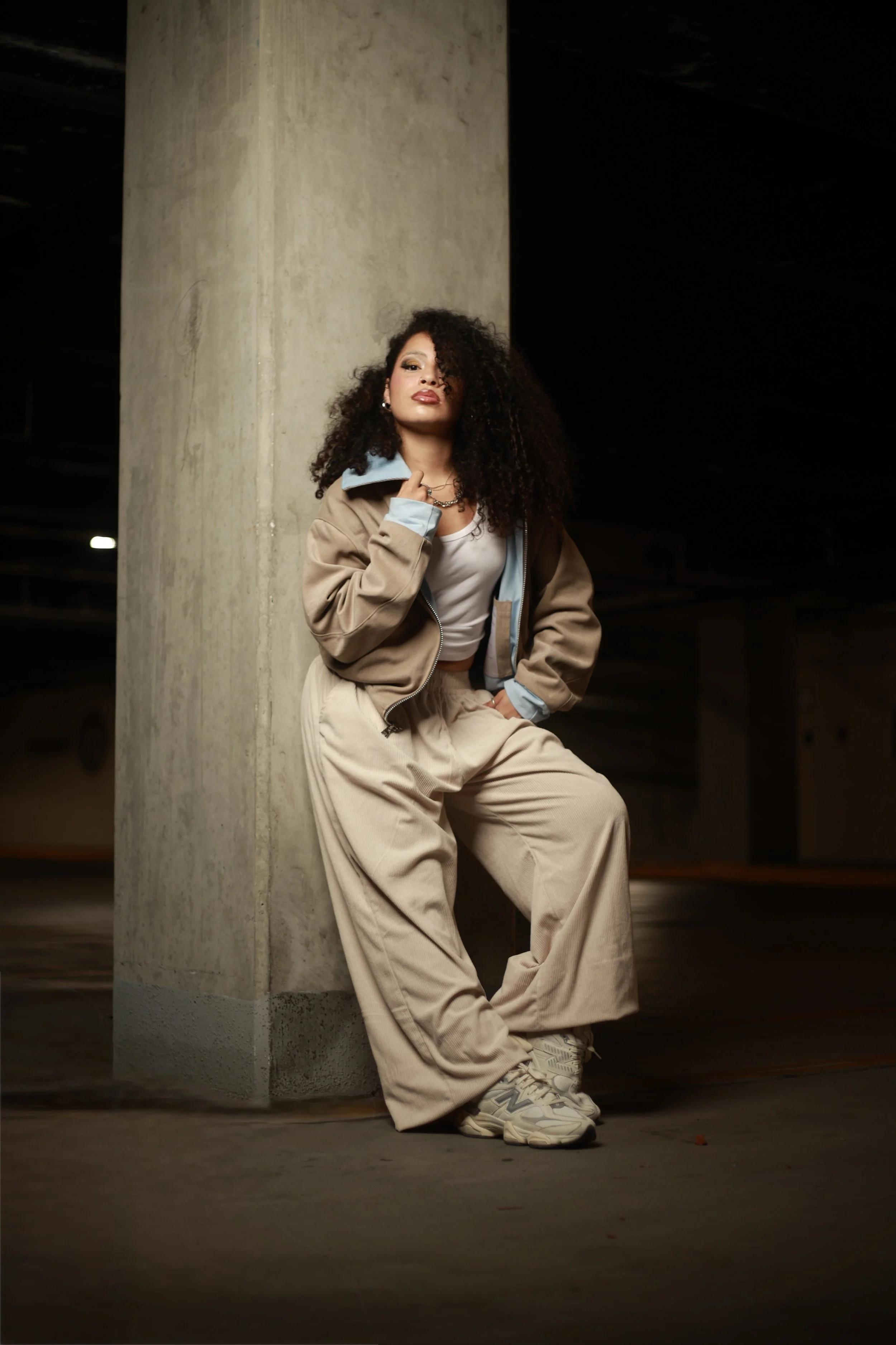 A woman with curly hair leaning against a concrete pillar in a dimly lit parking garage, wearing beige sweatpants, a beige jacket with a blue lining, a white top, and sneakers.