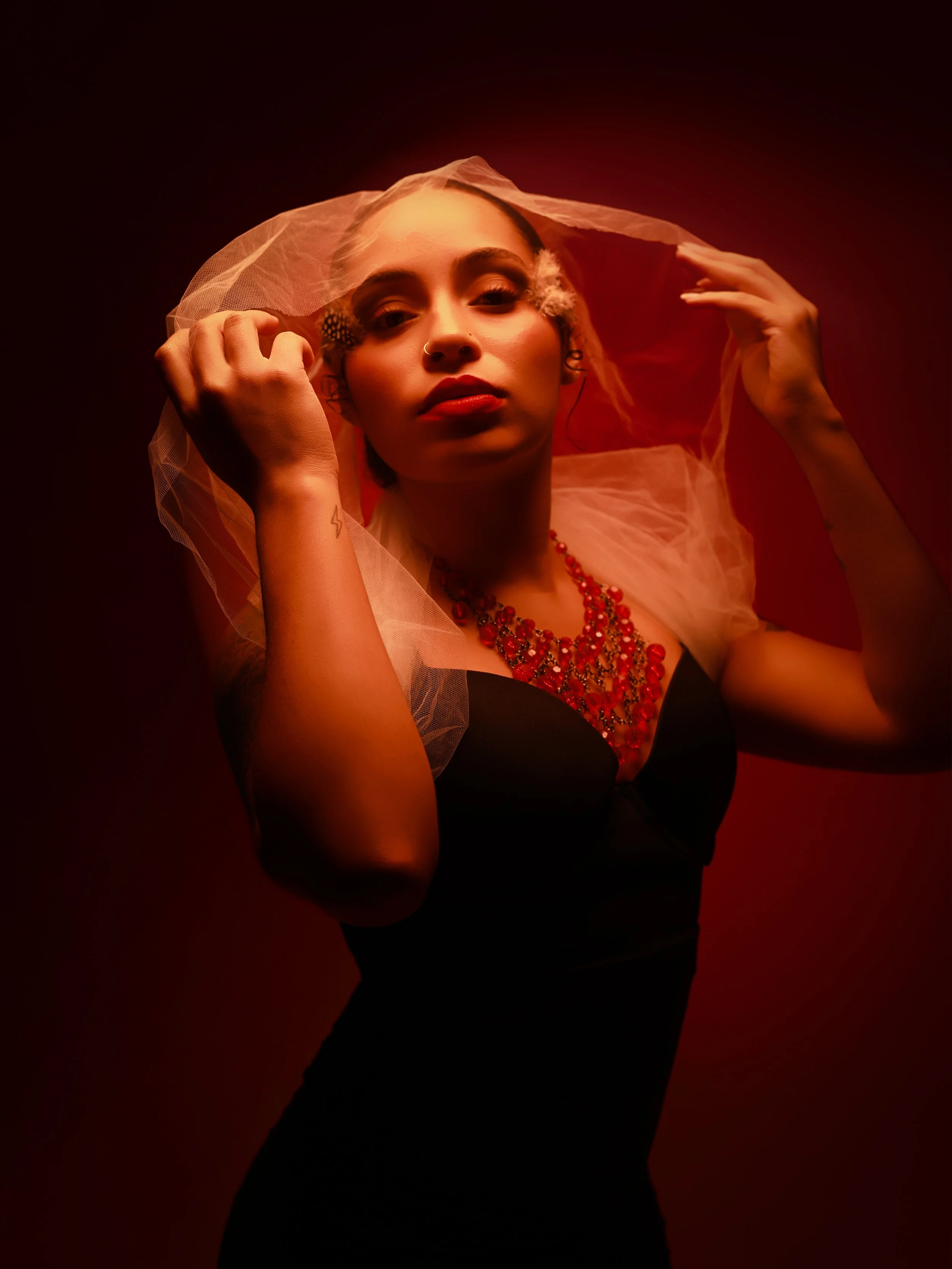 A woman with makeup, red lipstick, and a nose piercing holding a sheer white veil over her head, wearing lipstick, a red beaded necklace, and a tattoo on her wrist, against a red background.