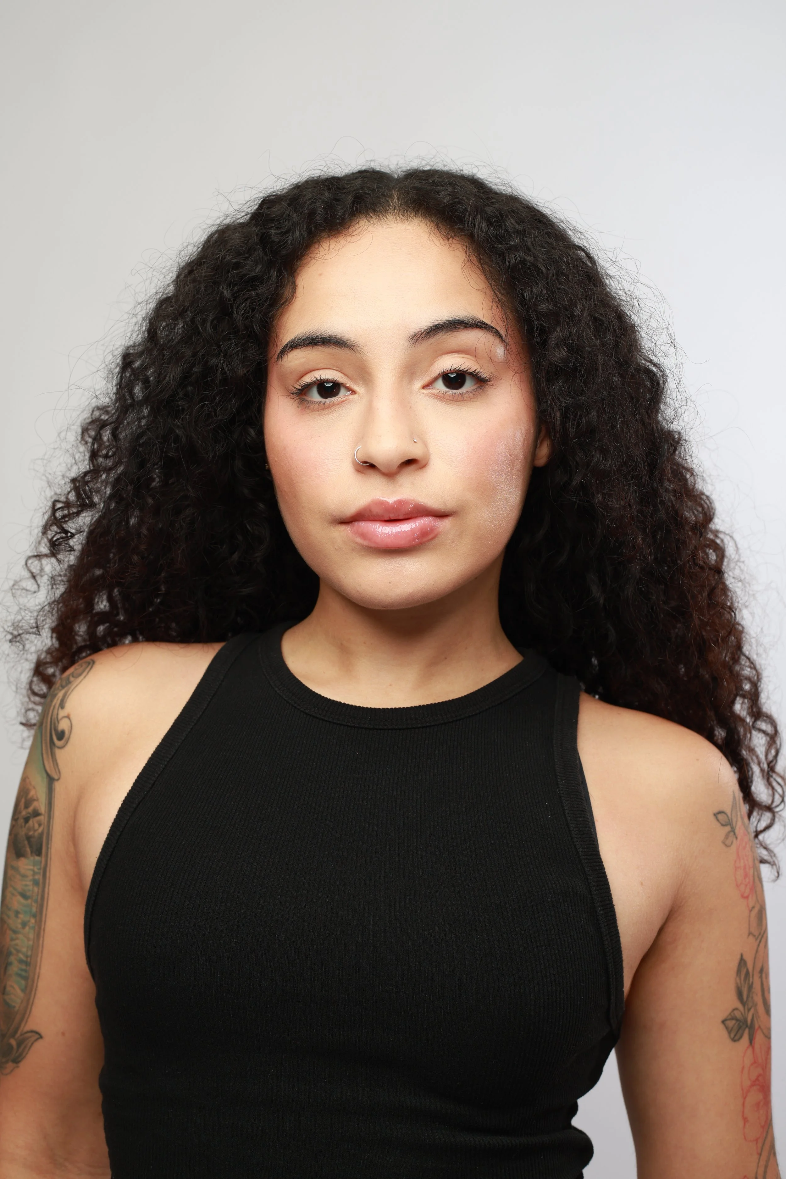 Close-up of a young woman with curly dark hair and tattoos, wearing a black sleeveless top, against a plain light background.