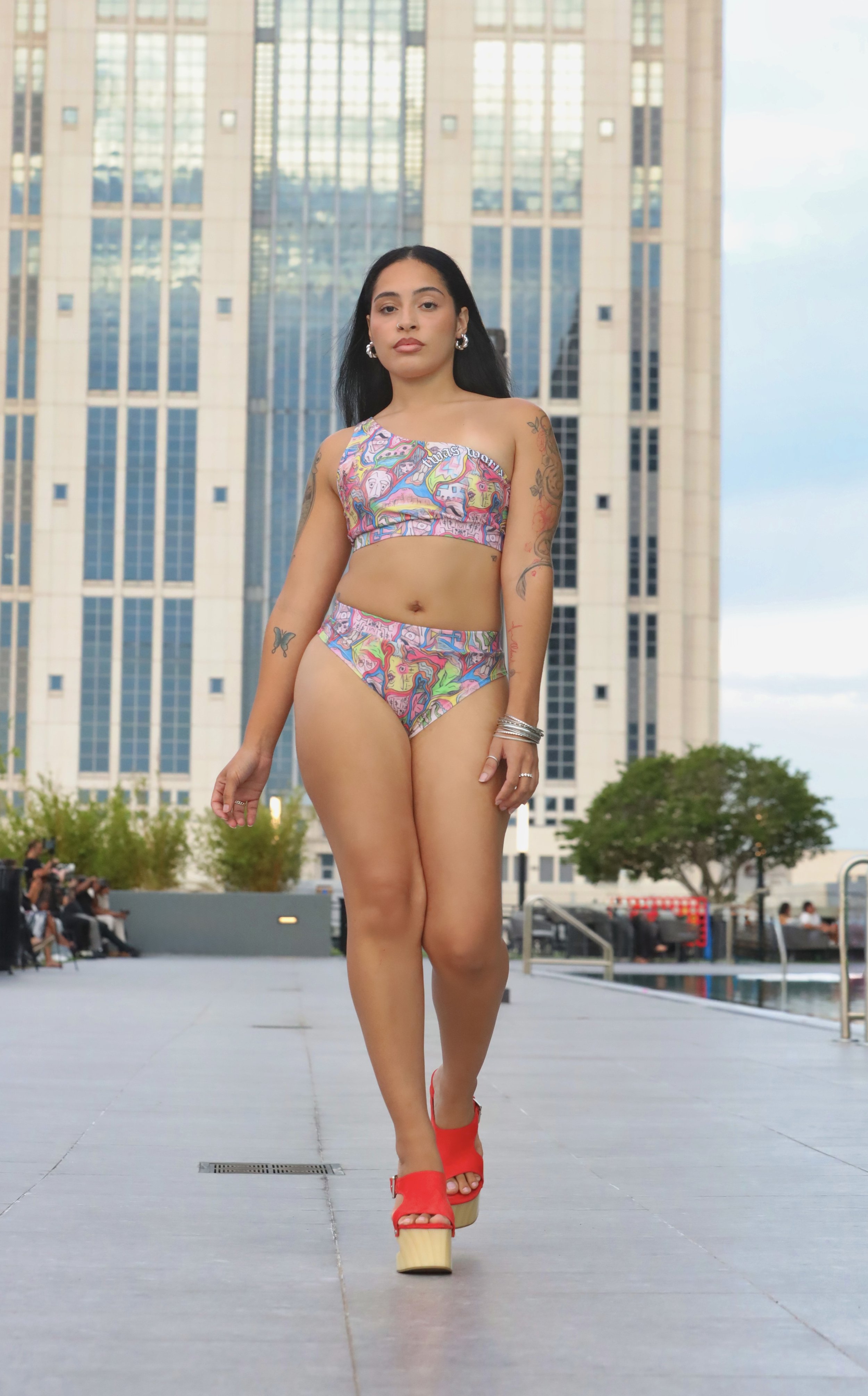A woman walking outdoors on a runway in front of tall buildings during daytime, wearing a colorful matching bikini and platform orange sandals.
