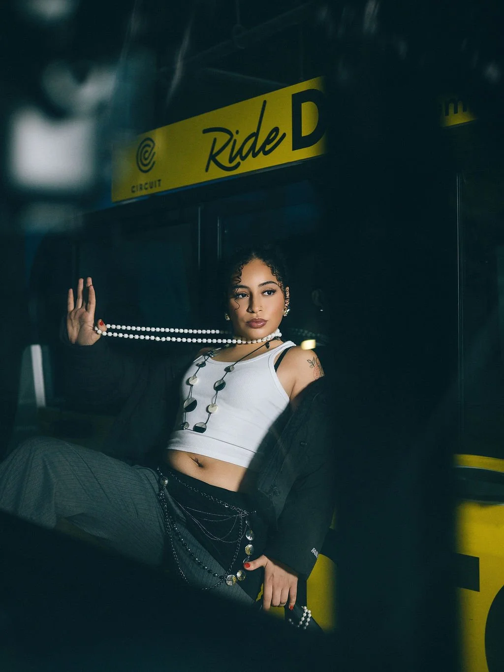 A young woman with curly hair and tattoos wearing a white crop top, jewelry, and a black jacket draped over her shoulders, sitting in front of a yellow sign that reads 'Ride D' at a transit station.