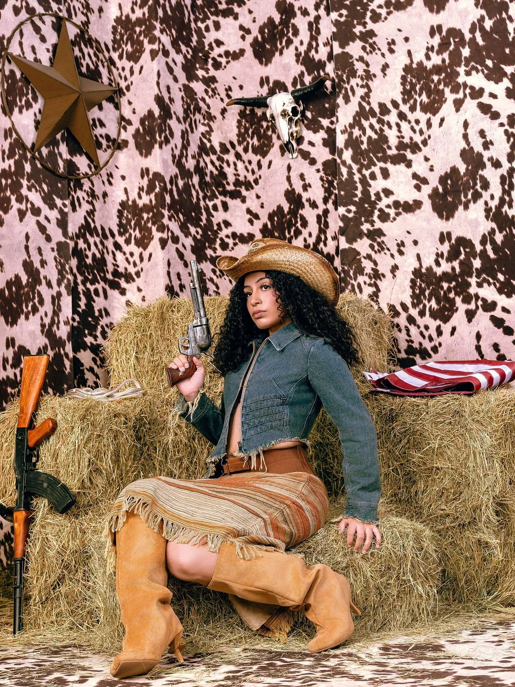 A woman dressed in cowboy attire sitting on hay bales in a western-themed setting. She is wearing a cowboy hat, a denim jacket, a skirt with fringes, and tan boots, and is holding a revolver. The background features a cow-print pattern on the wall, a star decoration, and a bull skull.