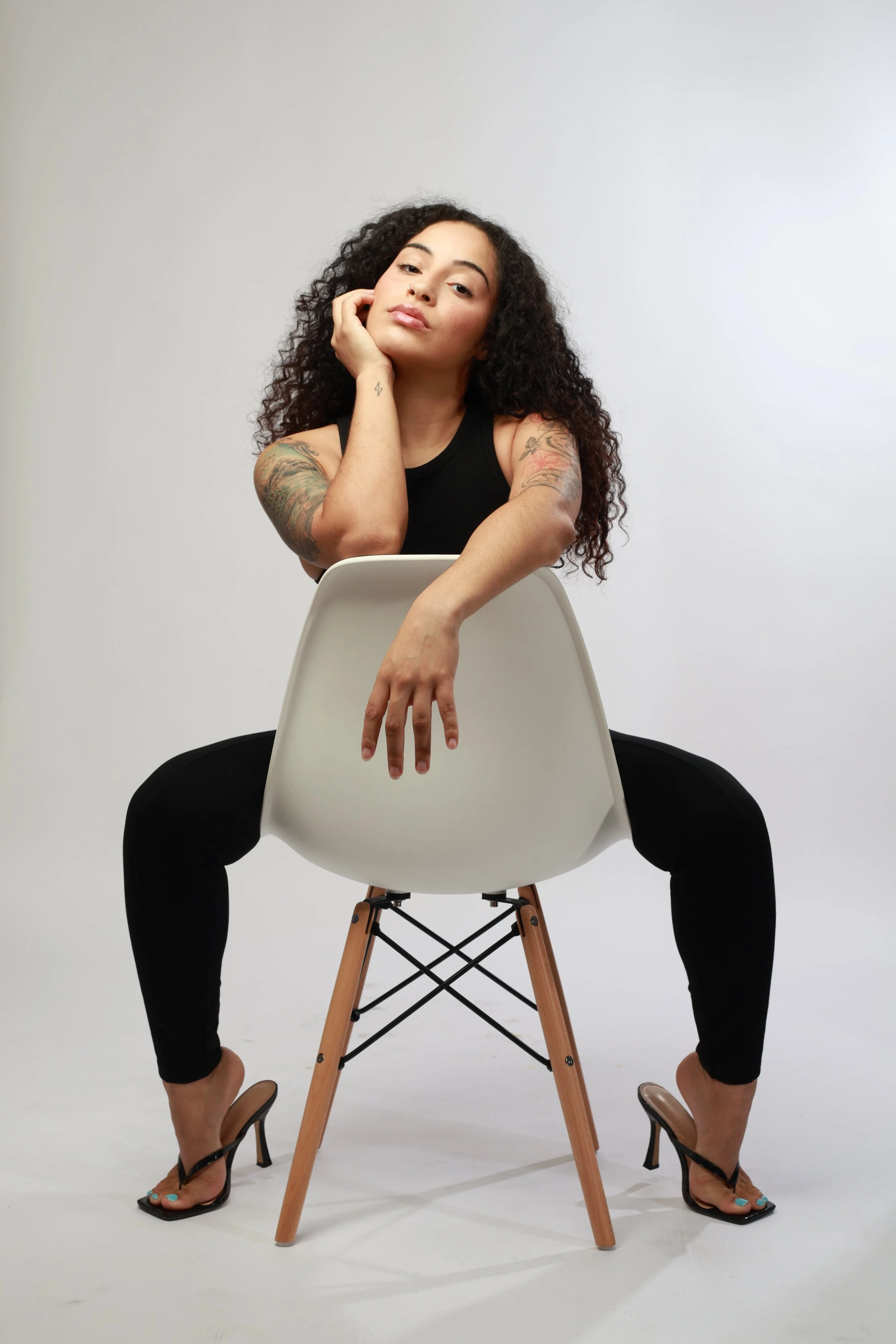 A woman with curly hair and tattoos on her arms sits on a white chair, resting her head on her hand, looking at the camera. She wears a black tank top, black pants, and high-heeled sandals. The image is captured in a studio with a plain white background.