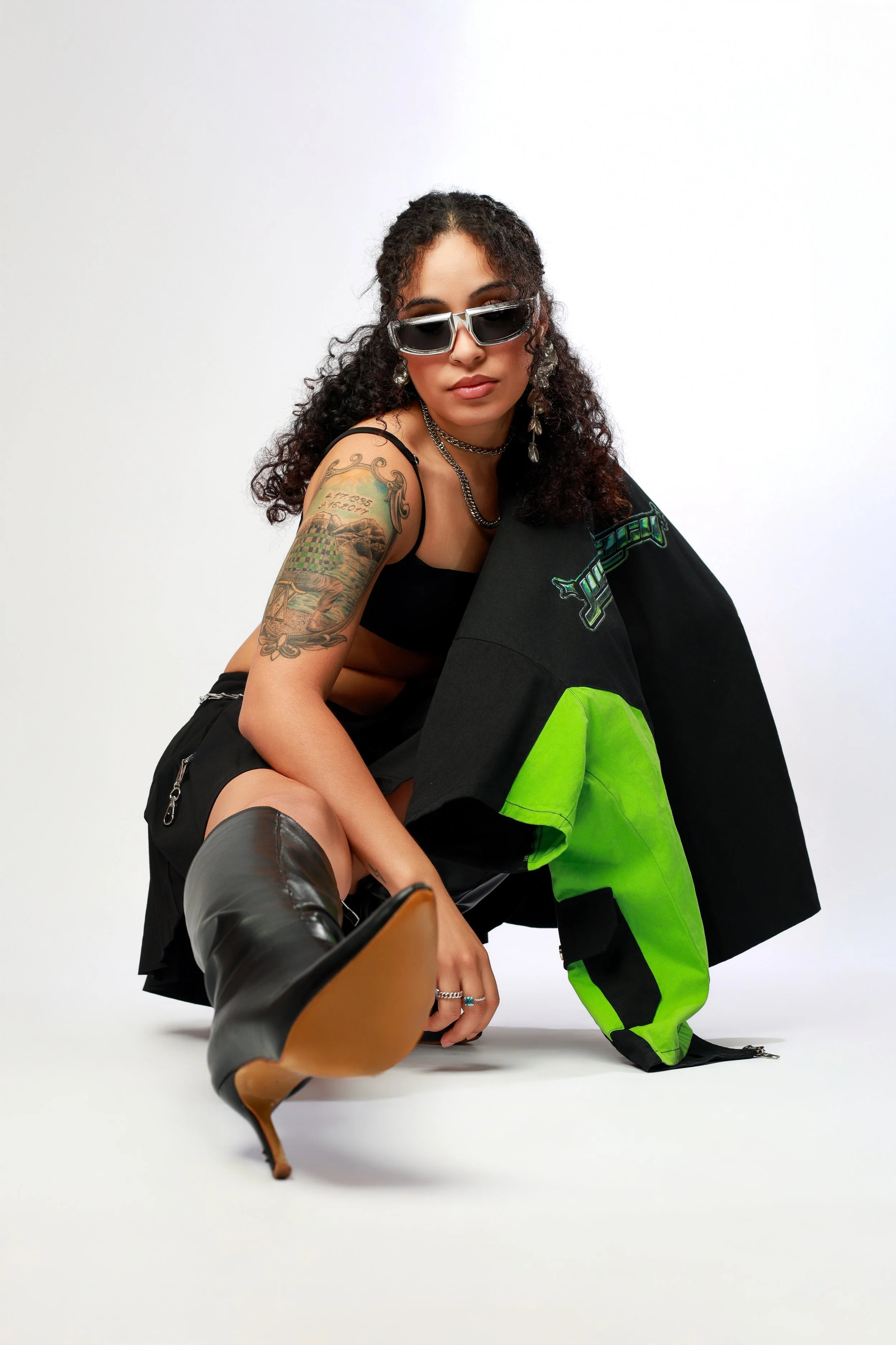 A woman with curly hair wearing sunglasses, a black top, and jewelry, sitting on the floor against a plain white background. She has a tattoo on her left arm and is dressed in dark pants and boots. She is leaning forward with one leg stretched out and the other bent, looking directly at the camera.