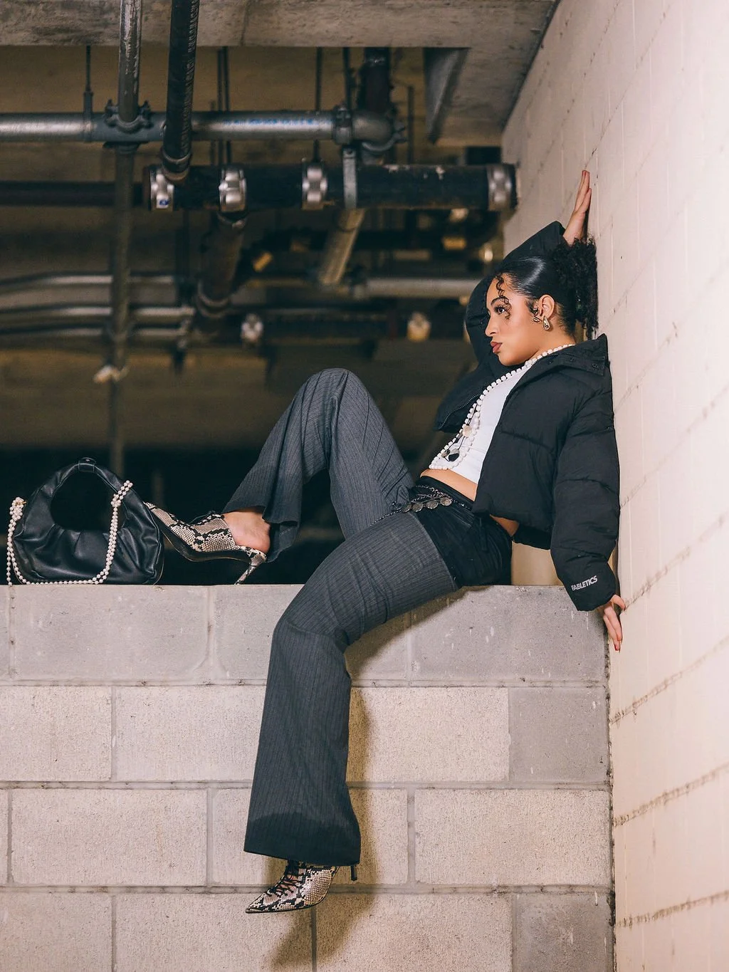 Fashionably dressed woman sitting on a concrete ledge in an industrial parking garage, leaning against a white brick wall with pipes overhead, accessorized with a pearl necklace, snake print shoes, and a black handbag.