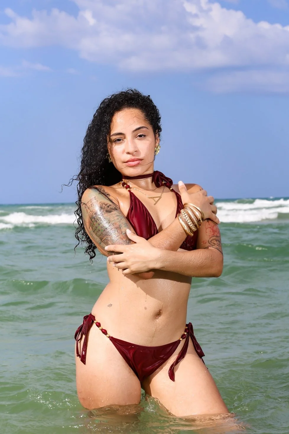 A woman with curly dark hair standing in the ocean, wearing a maroon bikini with gold accents, accessorized with jewelry and tattoos, under a partly cloudy sky.