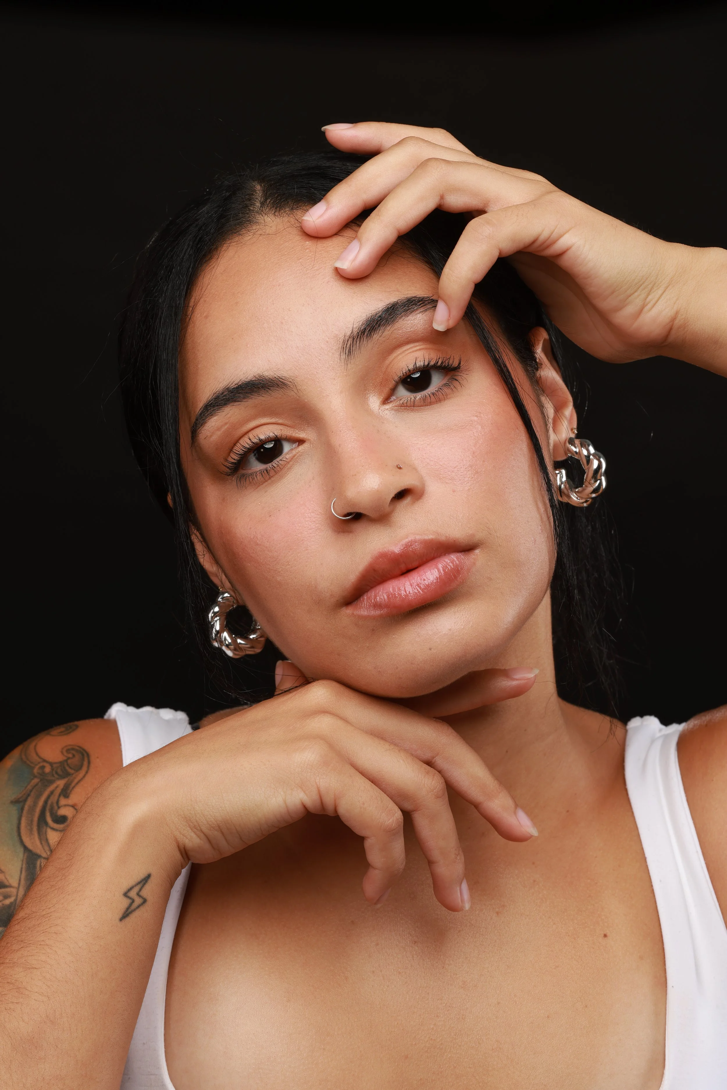 A young woman with dark hair, earrings, and tattoos is posing with her hand touching her forehead and chin against a black background.