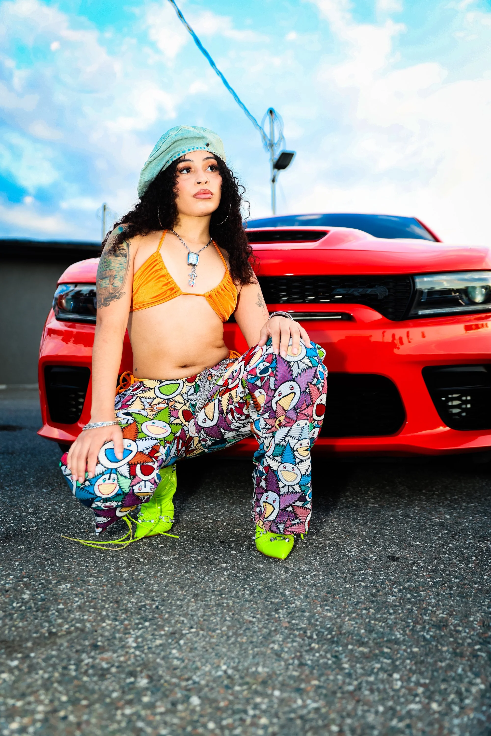 A woman with curly dark hair, wearing bright yellow boots, colorful patterned pants, a orange crop top, a necklace, and a mint green beret, is squatting in front of a red sports car on a parking lot with a cloudy blue sky in the background.