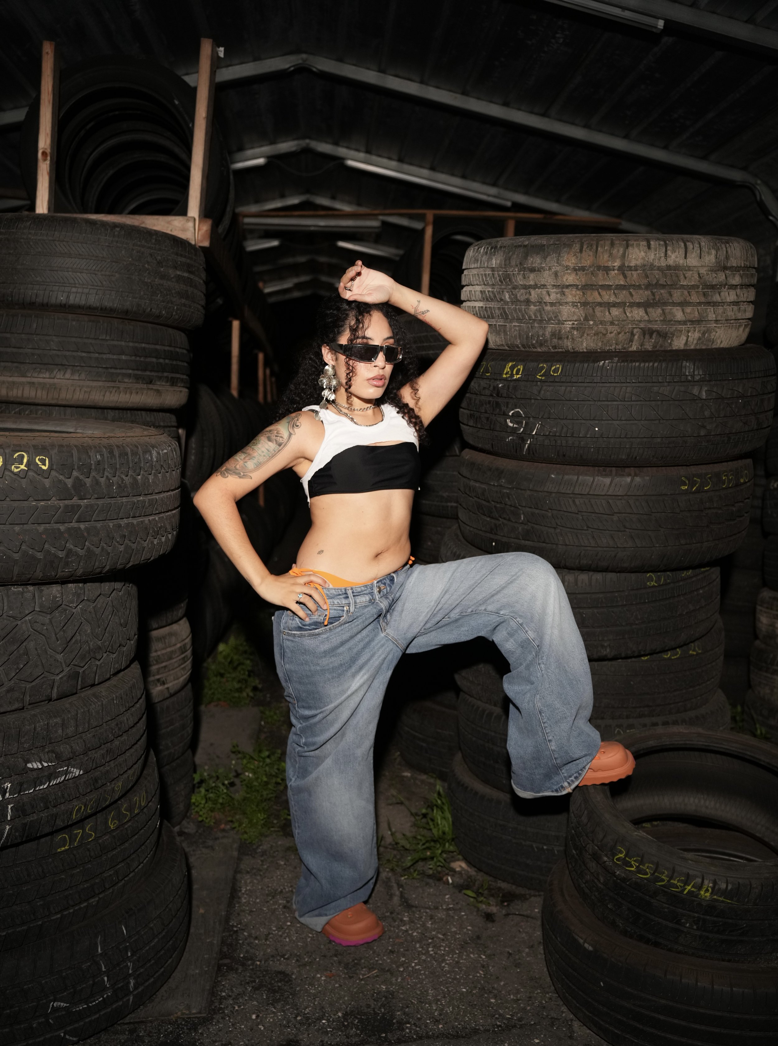 A woman with tattoos, sunglasses, and large earrings posing between stacks of tires in a warehouse or tire storage area.
