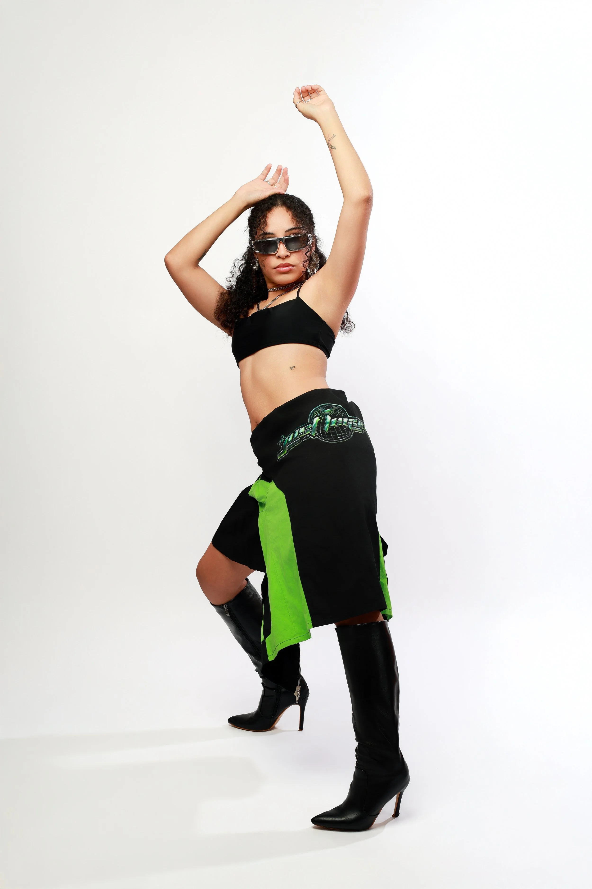 A woman with curly hair wearing sunglasses, a black crop top, black and neon green shorts, and black high-heeled boots, posing with one arm raised and the other covering her head against a plain white background.