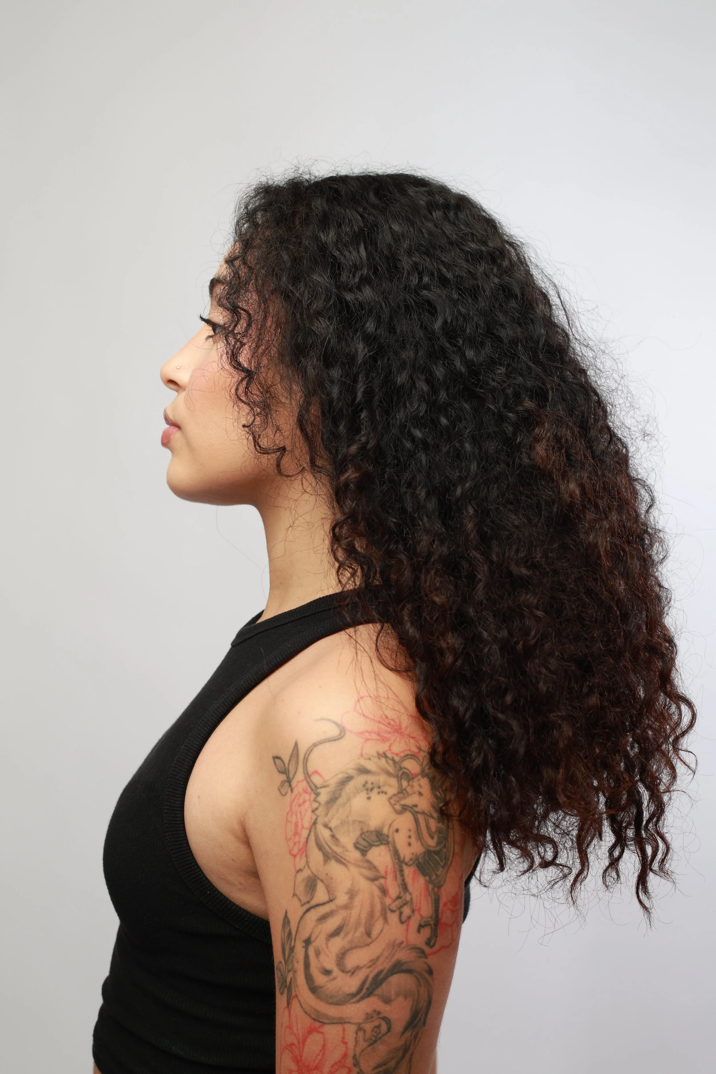 Side profile of a woman with dark curly hair, wearing a black tank top with tattoos on her arm, against a plain light gray background.